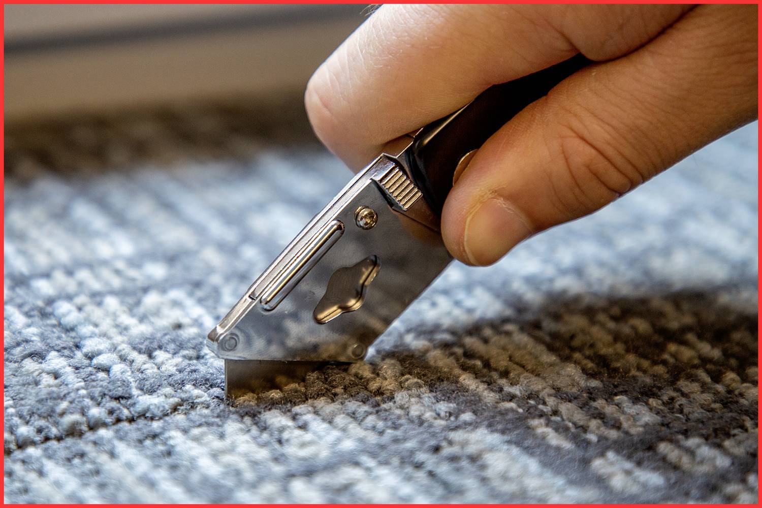 Eine Hand schneidet mit einem Teppichmesser einen Teppich. Das Messer hat eine silberne Klinge, und der Teppich ist graublau gemustert.