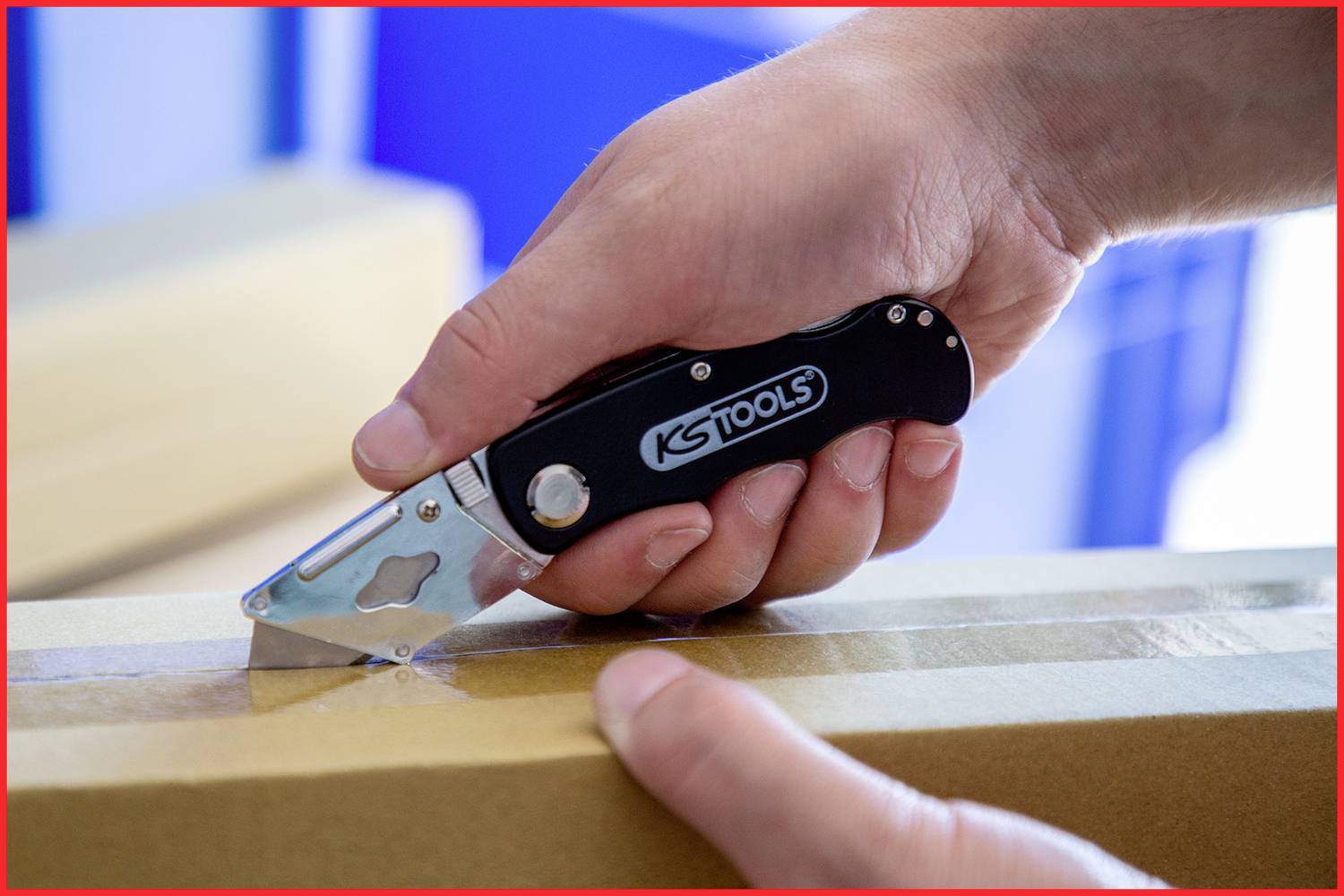 Hand hält ein Cuttermesser der Marke 'KS Tools' und schneidet Klebeband auf einem Karton. Hauptfokus liegt auf der Benutzung des Werkzeugs.