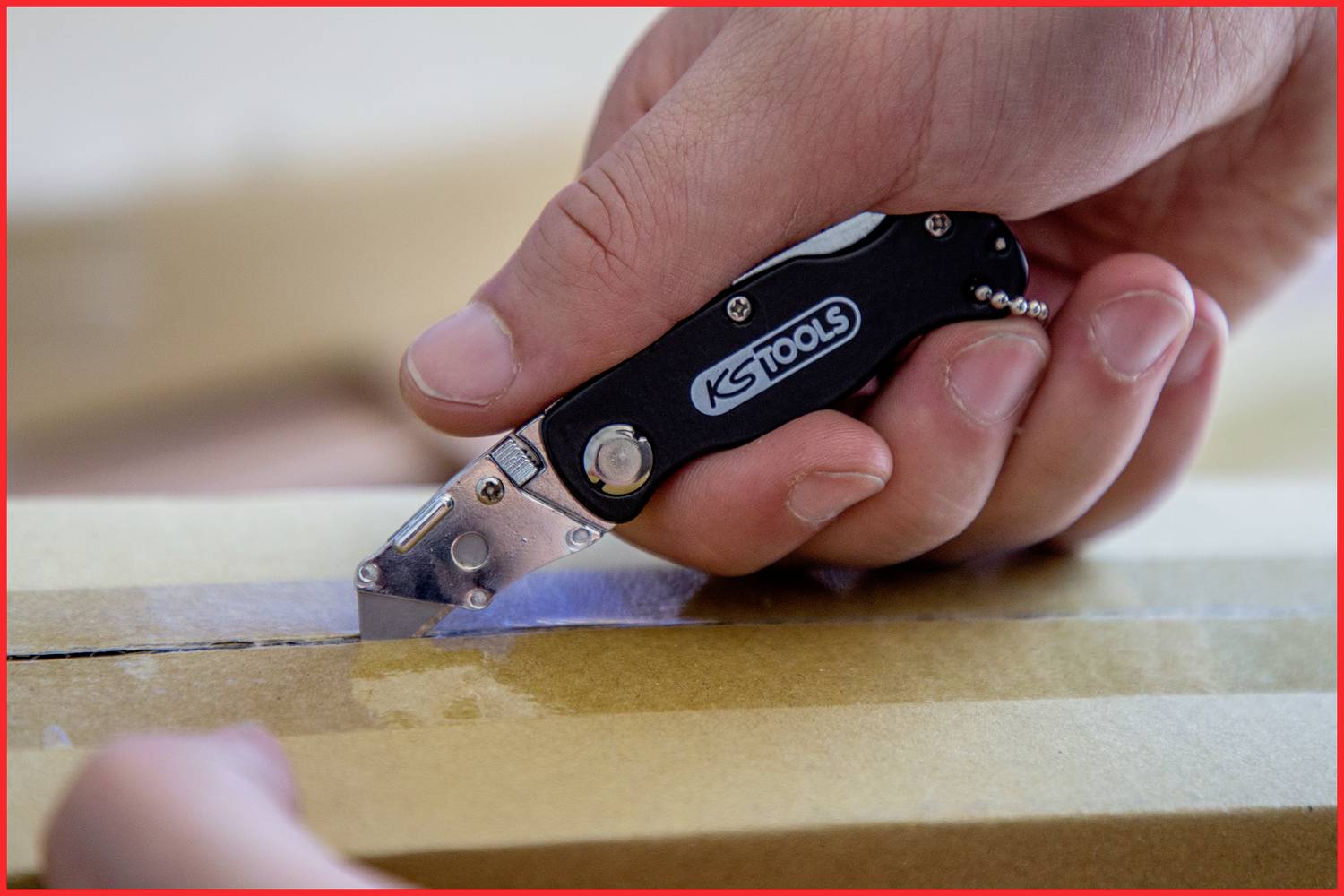 Eine Hand verwendet ein schwarzes Klappmesser, um Klebeband auf einem Pappkarton zu durchschneiden.