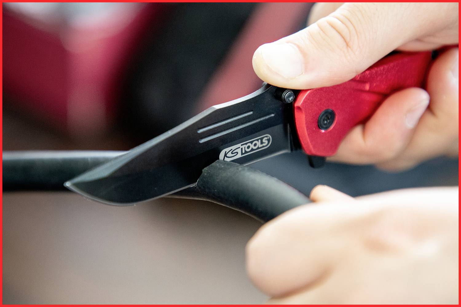 Eine Hand hält ein rotes Klappmesser und schneidet durch ein schwarzes Kabel oder Schlauch. Das Messer hat die Aufschrift 'KS TOOLS'.