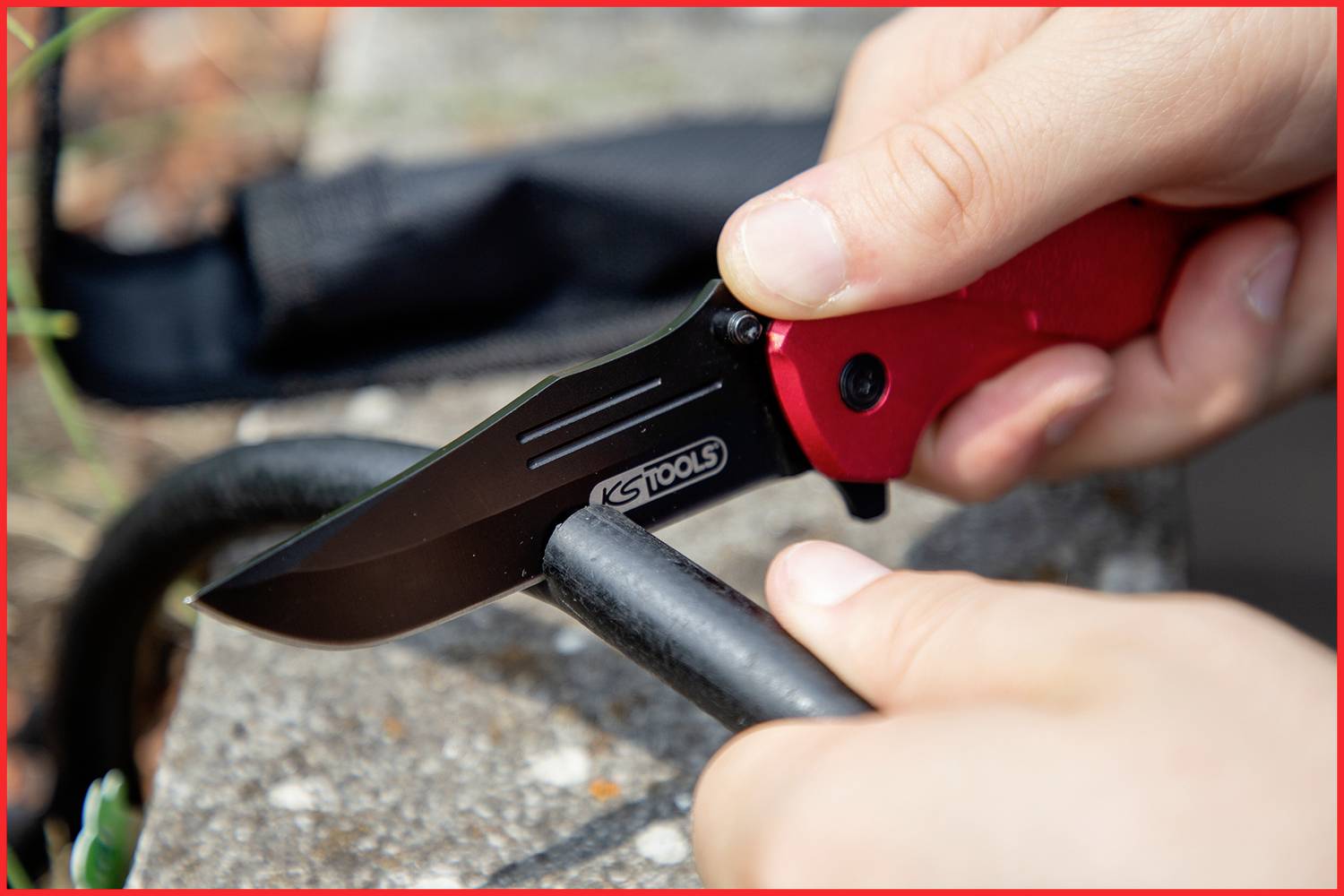 Person schneidet ein schwarzes Kabel mit einem roten Klappmesser auf einer Steinoberfläche. Nahaufnahme der Hände und des Messers.