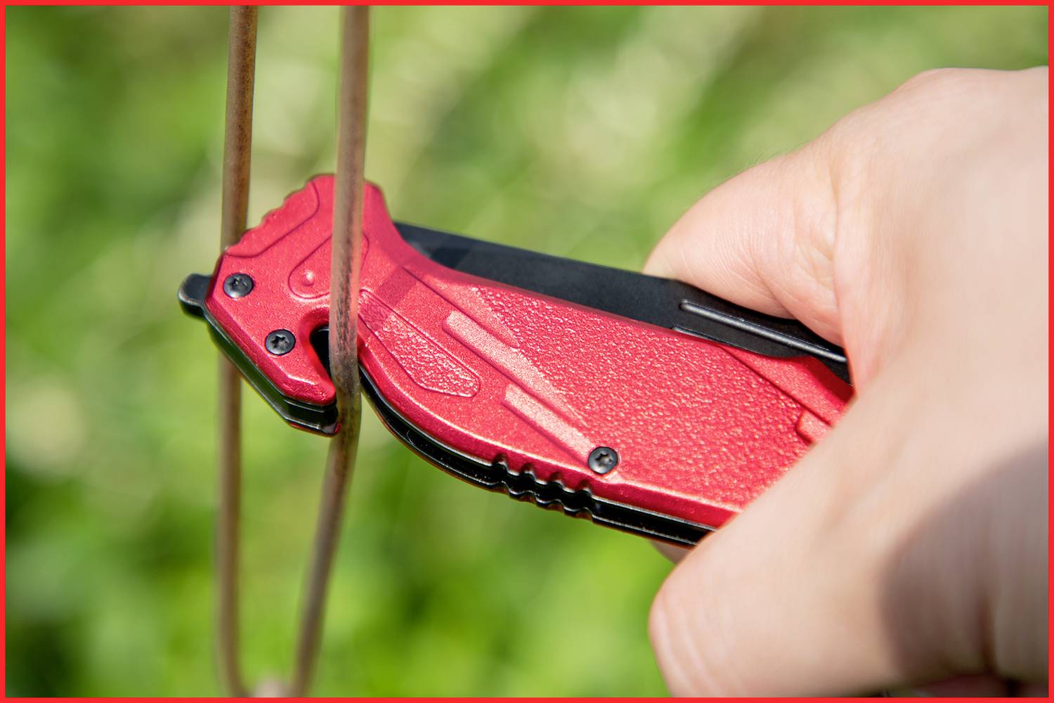 Eine Hand hält ein rotes Klappmesser, das ein dünnes Metallseil durchschneidet. Hintergrund aus unscharfem Grün.