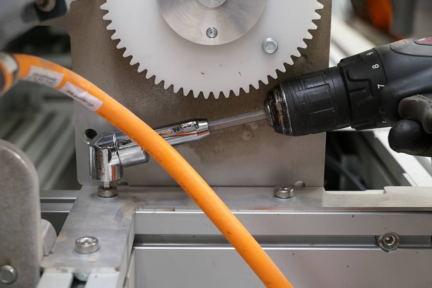 Eine Bohrmaschine wird benutzt, um ein Metallteil an einer Maschine zu befestigen. Im Hintergrund ist ein großes Zahnrad zu sehen.