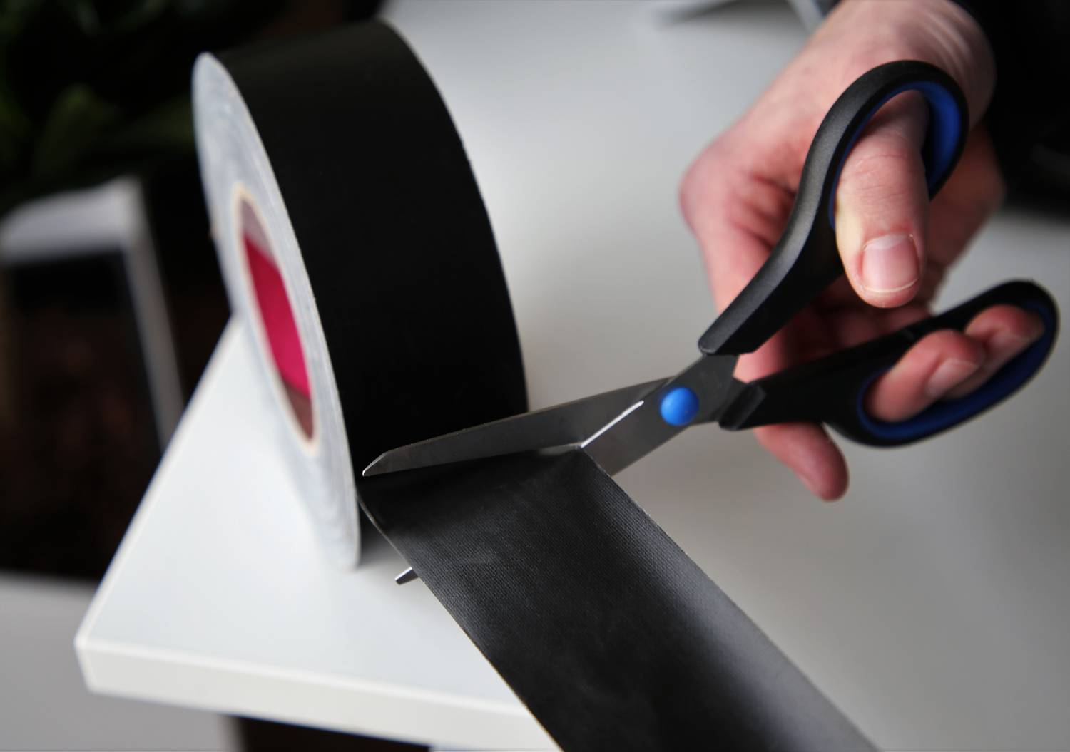 Eine Person schneidet ein Stück schwarzes Klebeband mit einer Schere. Das Band liegt auf einem weißen Tisch.