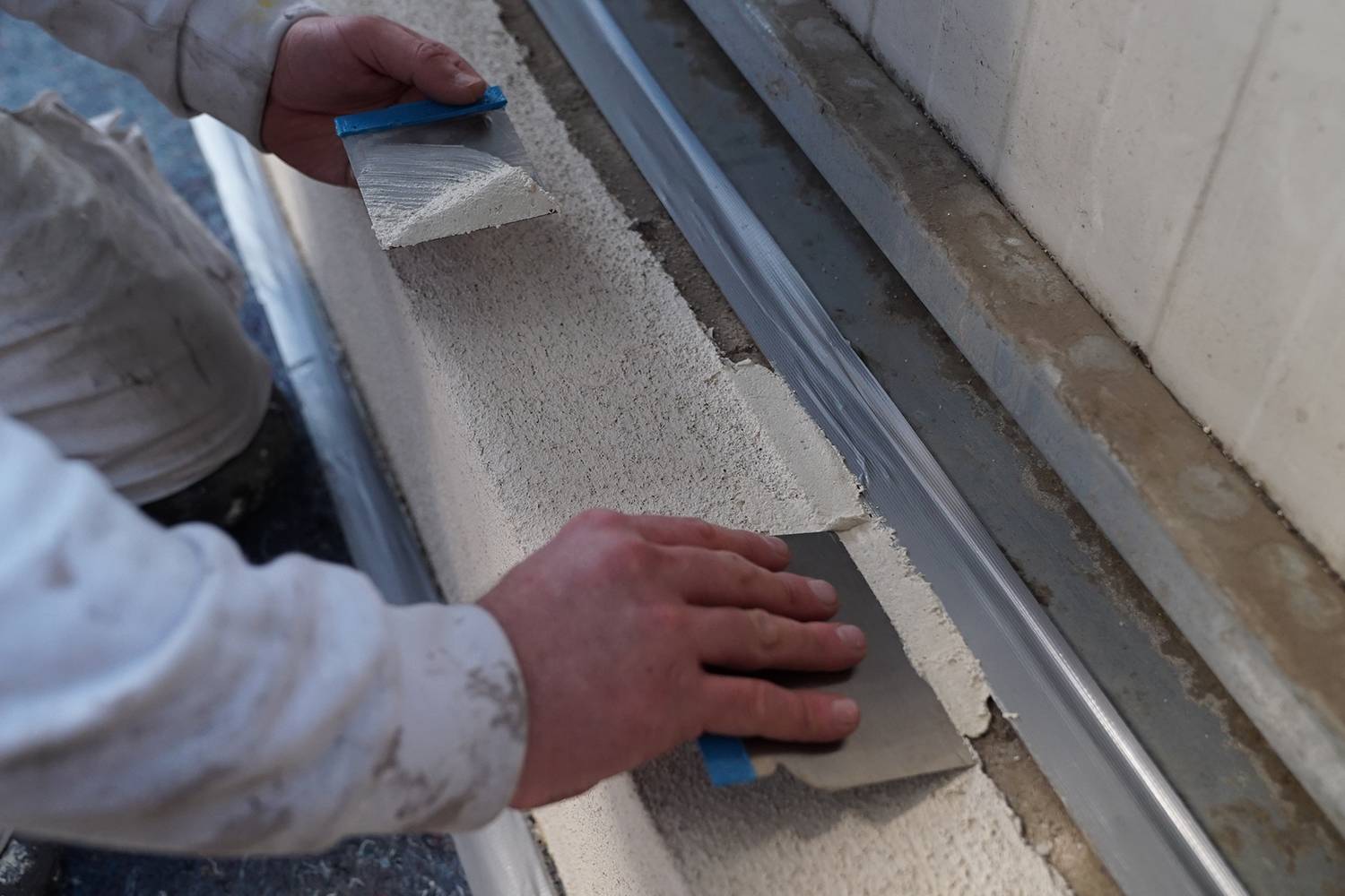 Eine Person trägt mit Spachteln weißen Mörtel auf eine Mauer auf. Fokus liegt auf den Händen und der Anwendungstechnik.