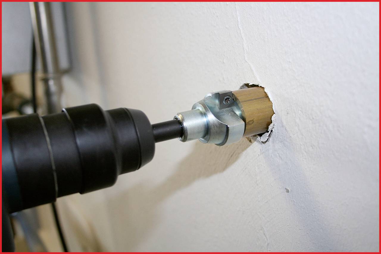 Eine Bohrmaschine mit Bohrkrone durchbohrt eine weiße Wand, um ein rundes Loch zu schaffen. Das Werkzeug ist schwarz und das Bohrzubehör silbern.