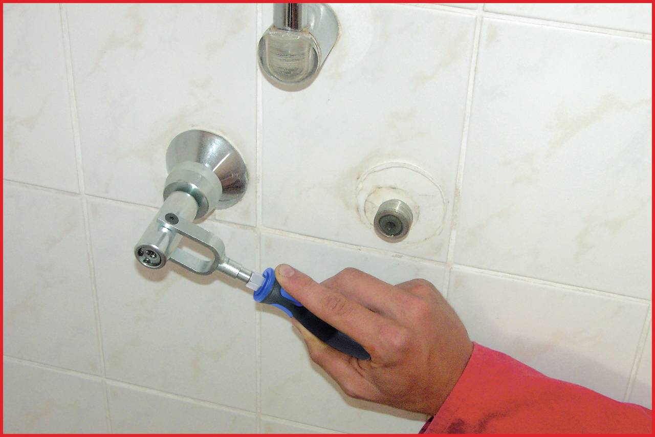Eine Hand hält einen blauen Schraubenschlüssel und dreht ein Ventil an einer Badezimmerwand, vermutlich zur Wasserregulierung.