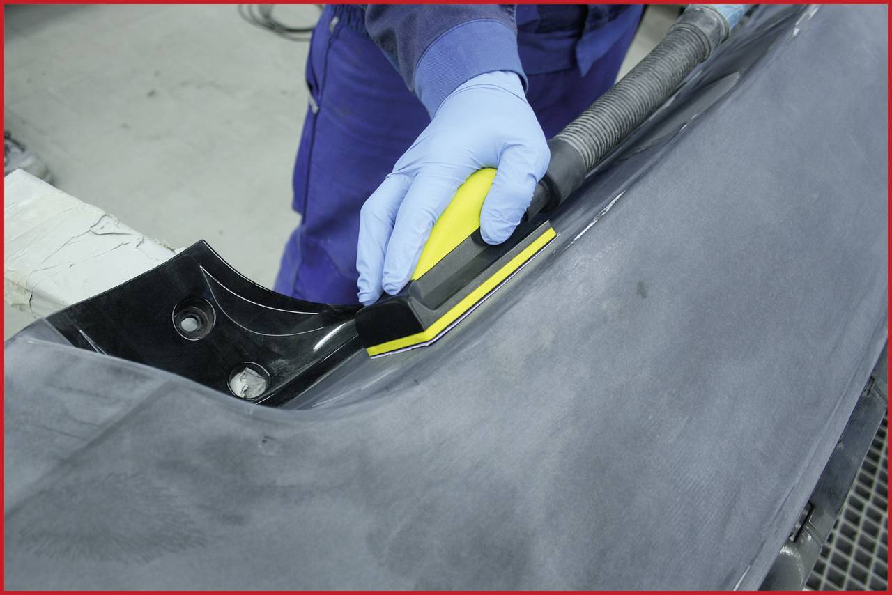 Eine Person trägt blaue Handschuhe und schleift eine schwarze Autoteilecke mit einem gelben Schleifwerkzeug in einer Werkstatt.