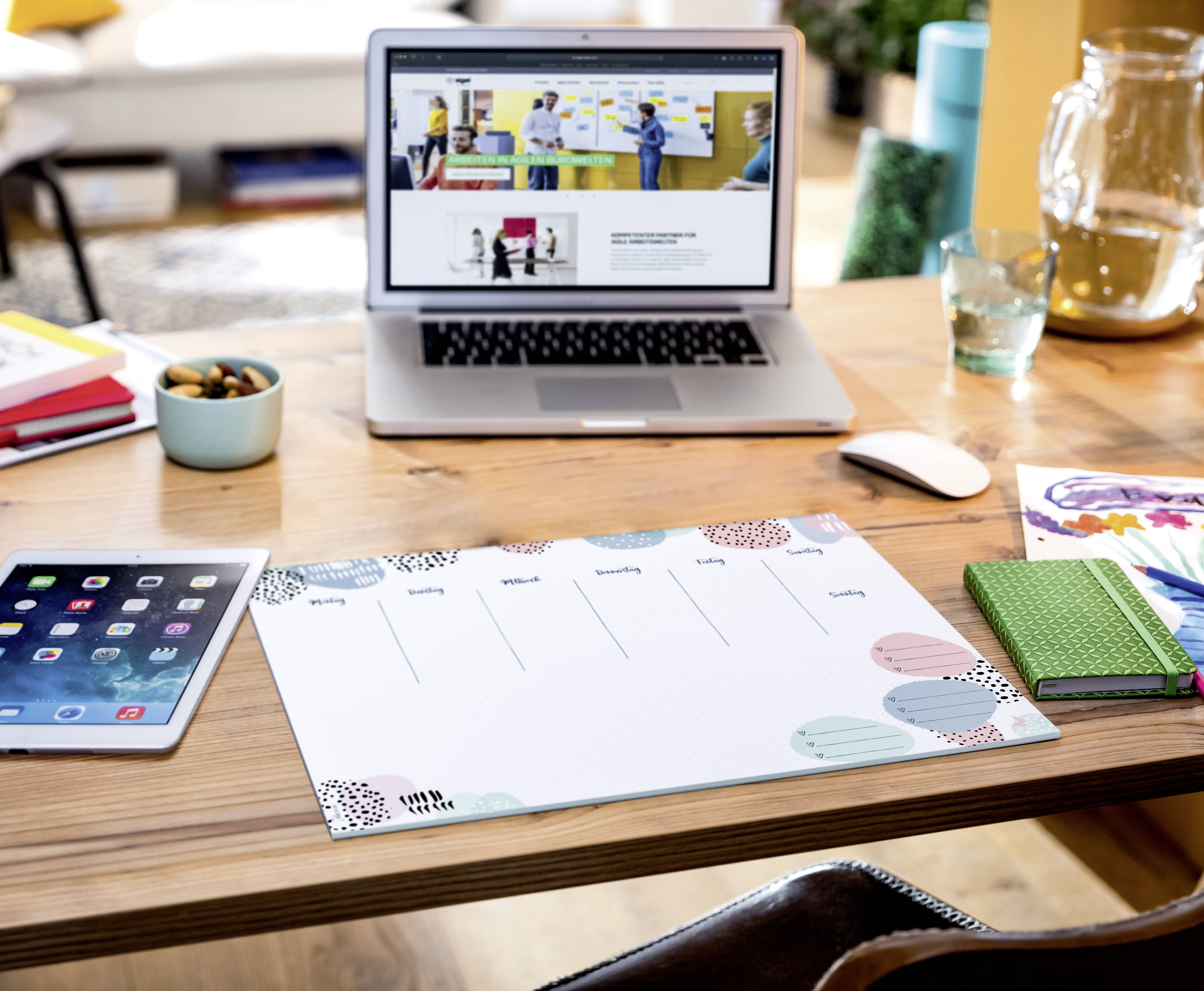 Schreibtisch mit Laptop, Tablet, Notizblock und Wochenplaner. Der Laptop zeigt eine Webseite mit Bildern von Menschen, die präsentieren.