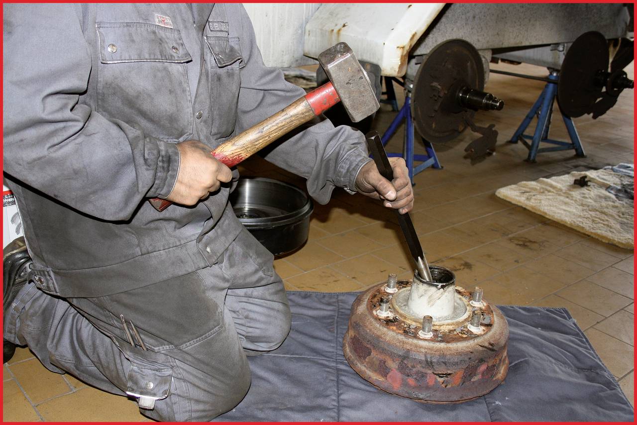 Eine Person in Arbeitskleidung repariert eine rostige Bremstrommel mit Hammer und Meißel in einer Werkstatt. Im Hintergrund sind Autoteile.