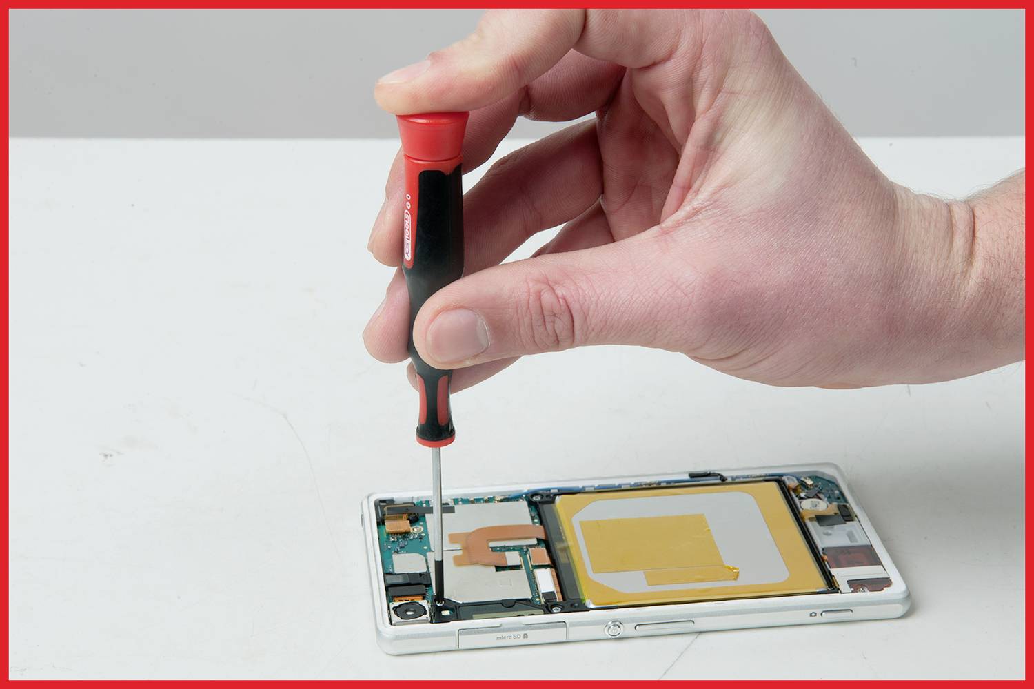 Nahaufnahme einer Hand, die mit einem Schraubendreher ein Smartphone öffnet. Der Innenaufbau zeigt Elektronikkomponenten.