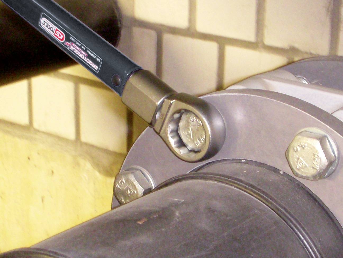 Ein Ratschenwerkzeug zieht eine große Schraube an einem Rohr fest. Im Hintergrund sind Fliesenwände sichtbar.