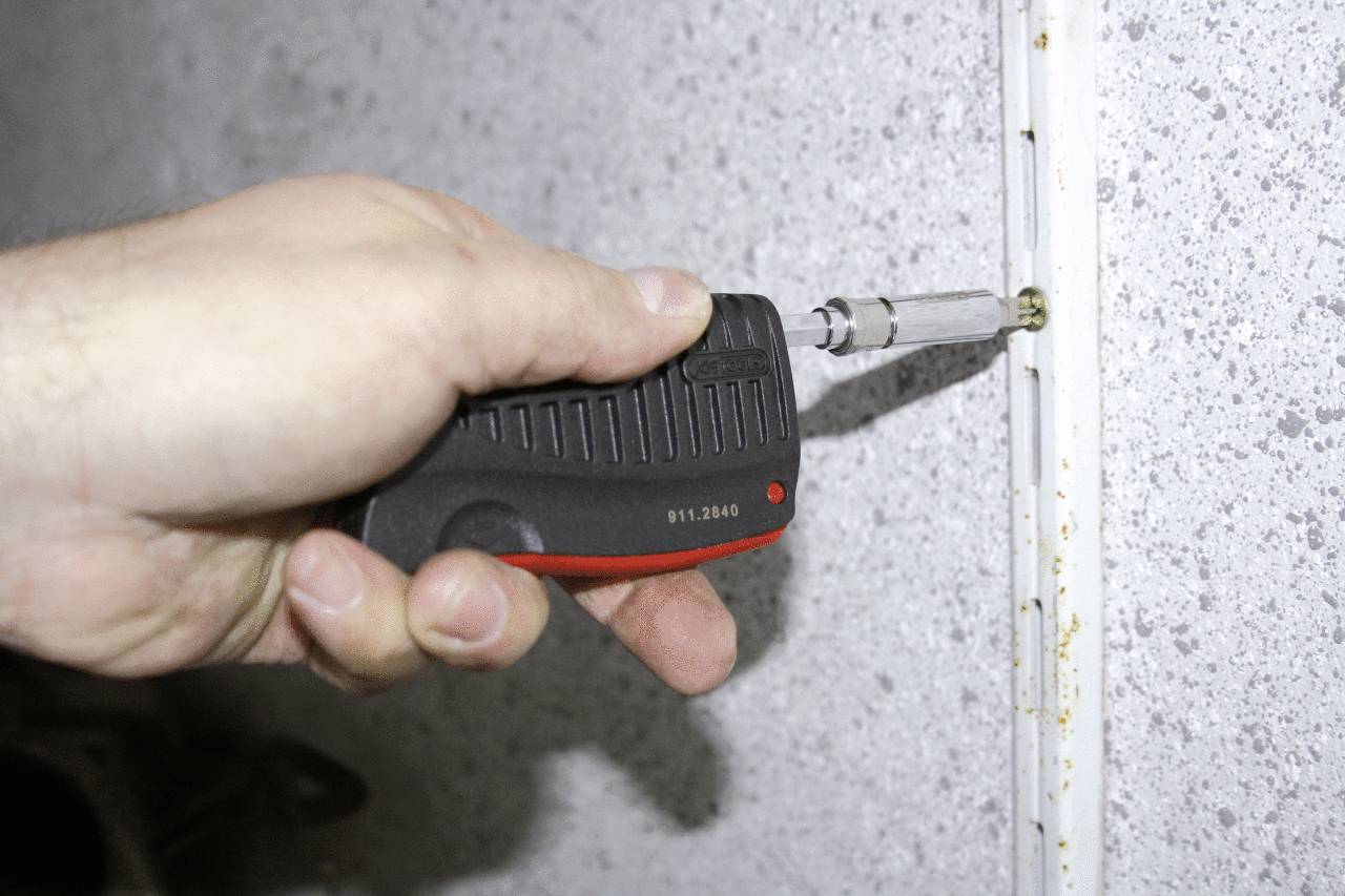 Eine Hand hält einen Schraubendreher und zieht eine Schraube in eine graue Wand fest, als Teil einer Montage- oder Reparaturarbeit.