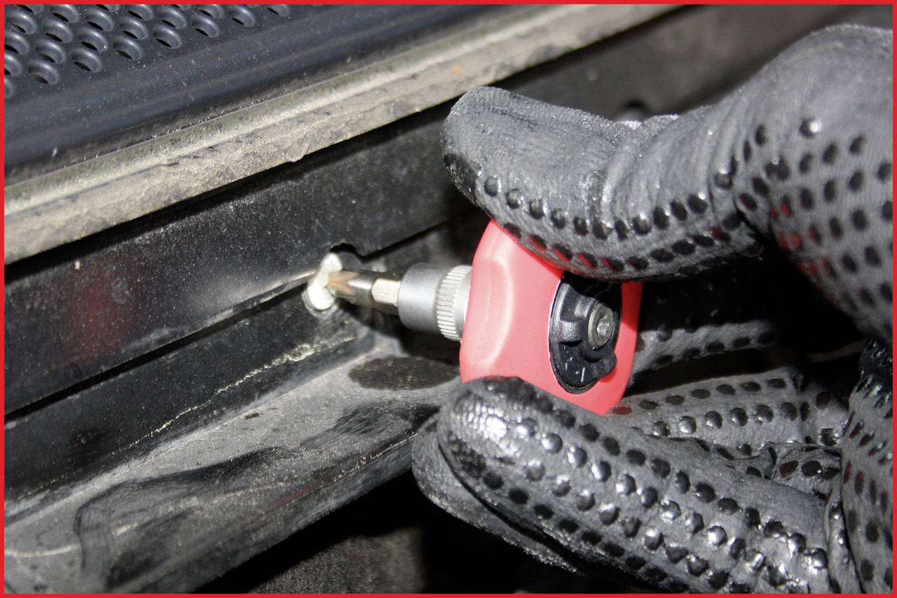 Nahaufnahme einer behandschuhten Hand, die mit einem Schraubendreher an einer schwarzen Oberfläche arbeitet.