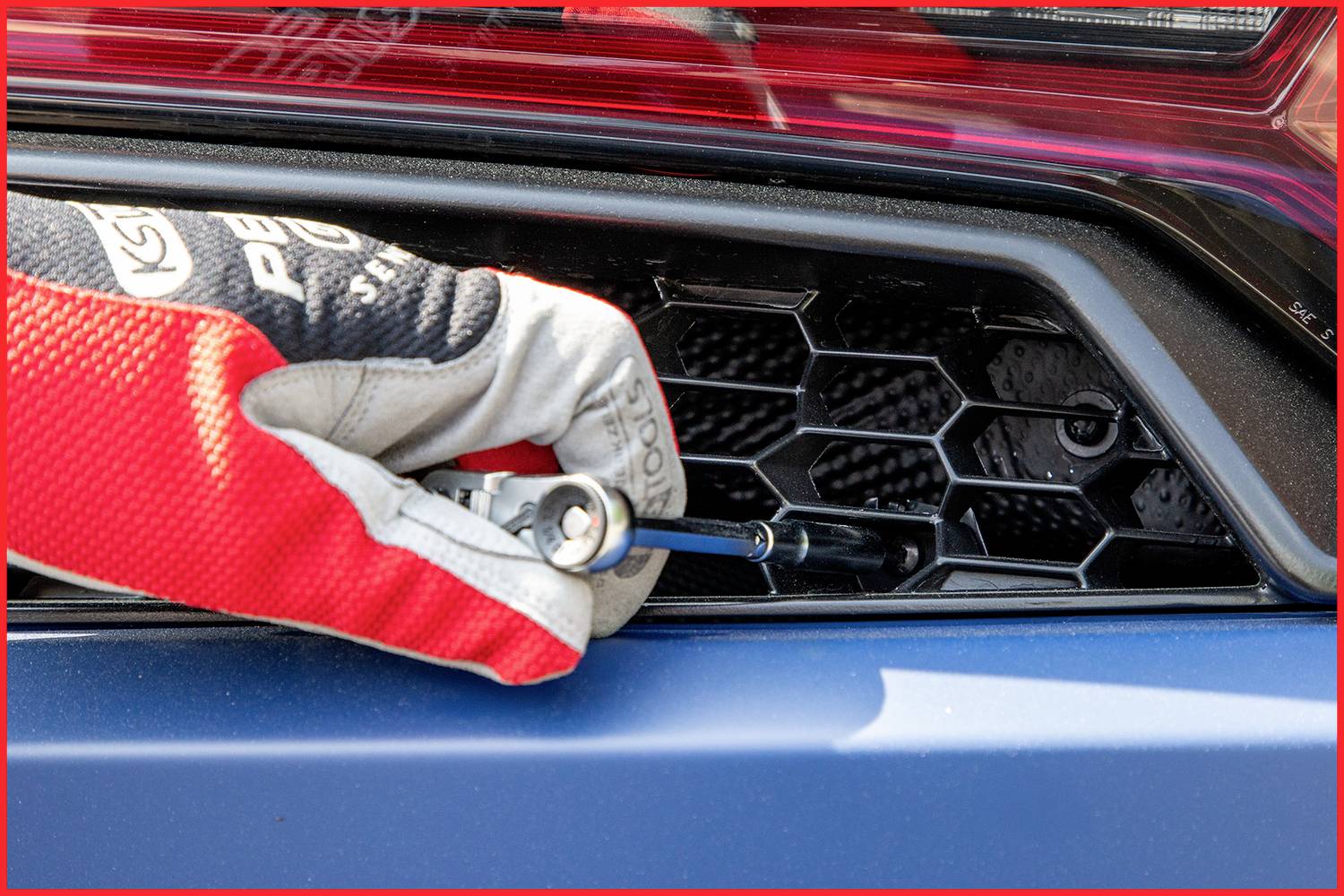 Eine Person mit einem Handschuh nutzt Werkzeug, um eine Wabengitterabdeckung an einem Fahrzeugheck zu befestigen oder zu entfernen.