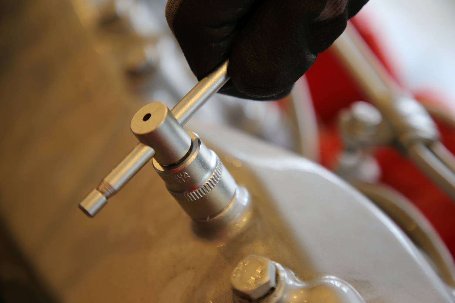 Hand mit Handschuh hält Werkzeug über Metallteil, das in eine Maschine eingebaut ist.