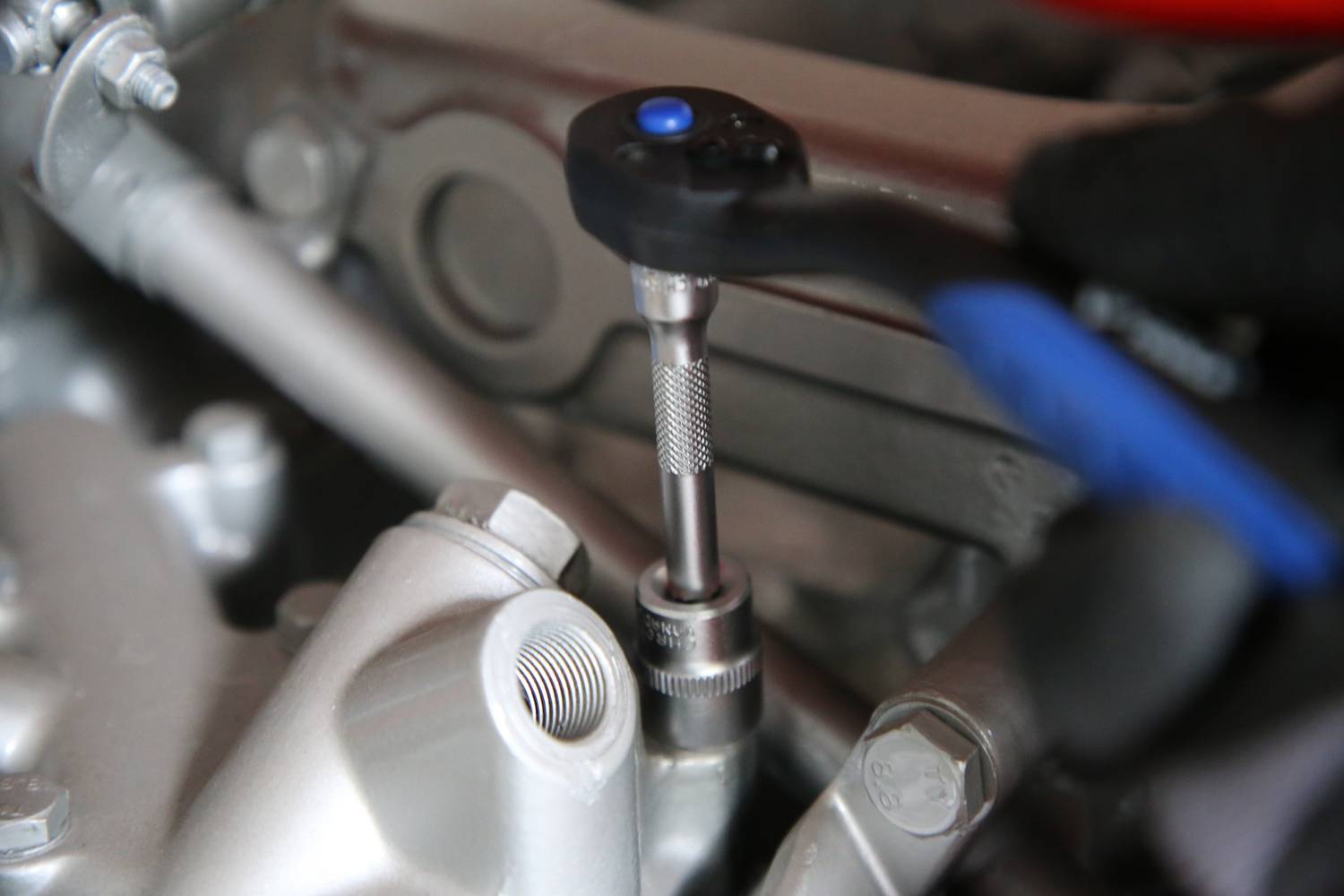 Eine Hand mit Handschuh dreht mit einem Ratschenwerkzeug eine Schraube im Motorraum eines Fahrzeugs fest.