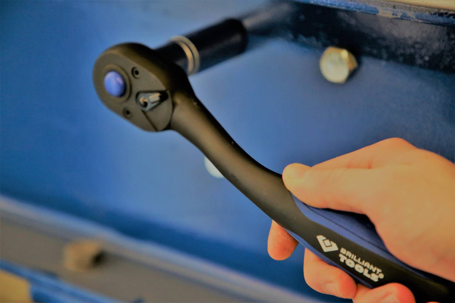 Eine Hand hält einen schwarzen Ratschen-Schlüssel, der an einer blauen Metalloberfläche arbeitet.