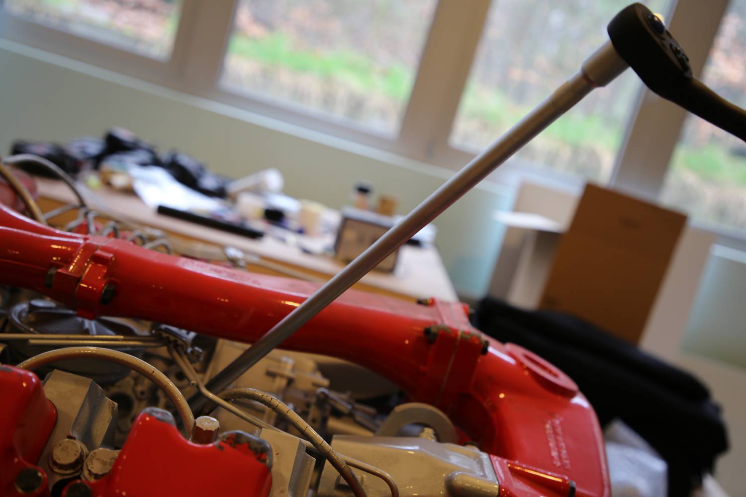 Ein roter Rennwagenmotor auf einem Tisch; Werkzeuge und Autoteile im Hintergrund; Fenster bieten Blick auf grüne Landschaft.