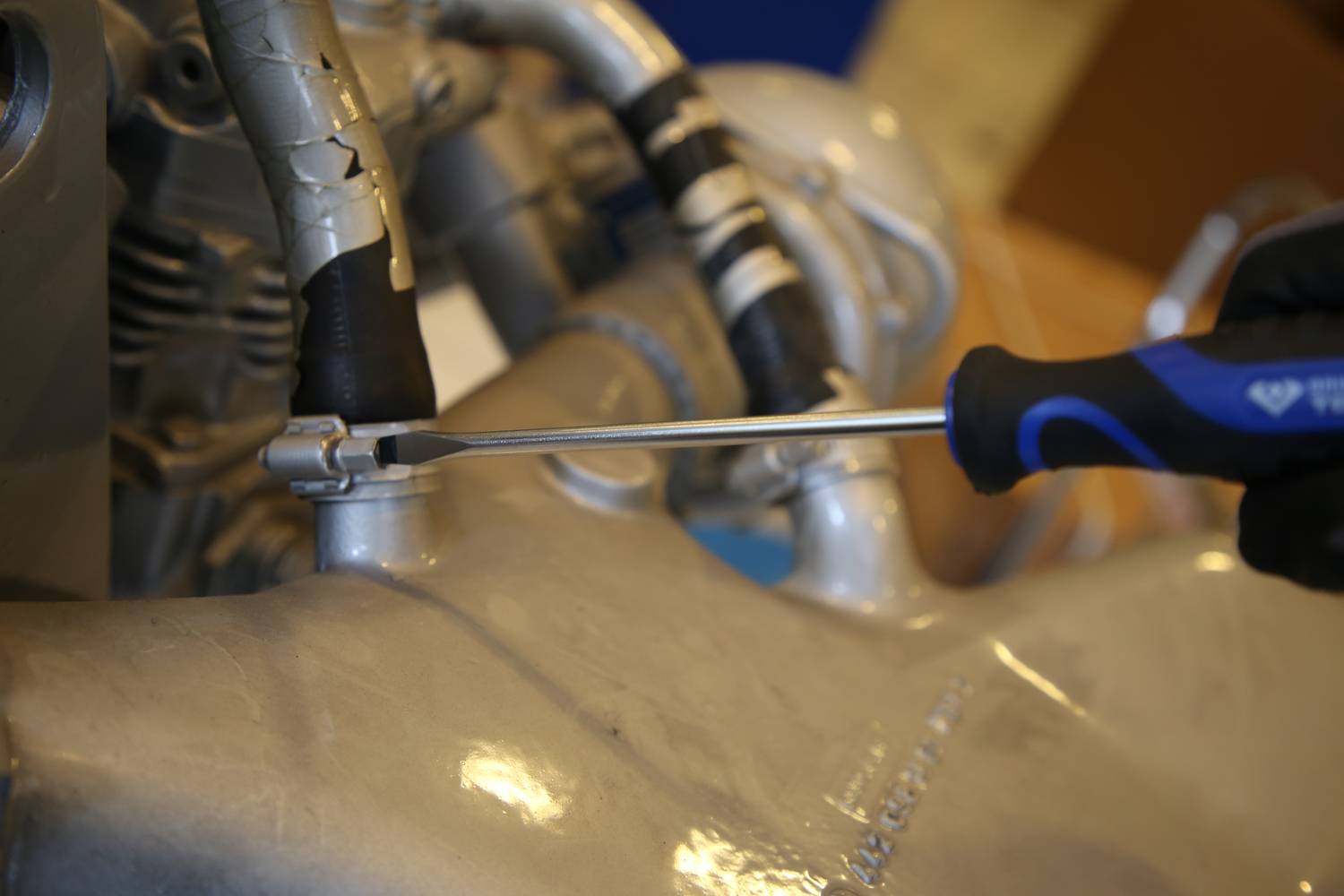 Eine Hand zieht mit einem Schraubendreher eine Schraube an einem Motorradmotor fest. Der Motor ist aus Metall und hat mehrere Schläuche.