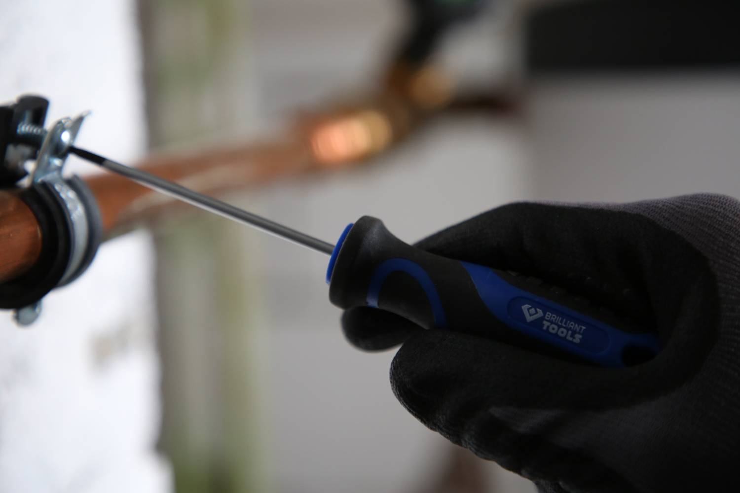 Hand in einem schwarzen Handschuh hält einen Schraubendreher und befestigt eine Schelle an einem Kupferrohr.