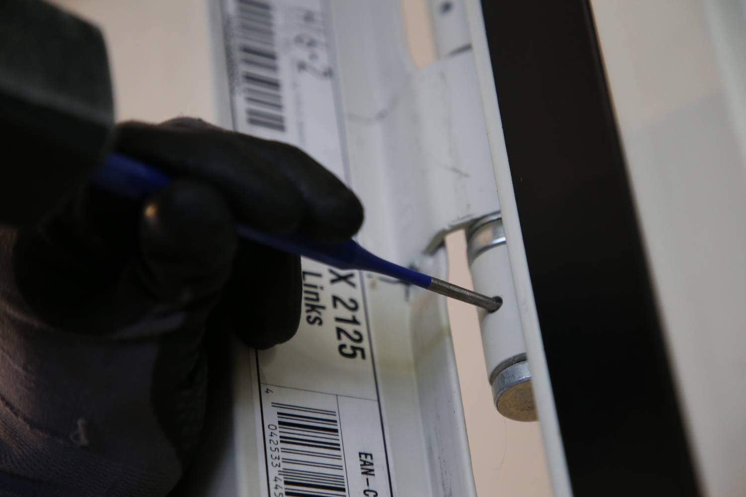 Eine Person mit einem schwarzen Handschuh zieht mit einem Werkzeug eine Schraube an einem Scharnier fest, neben einem Etikett mit Text.