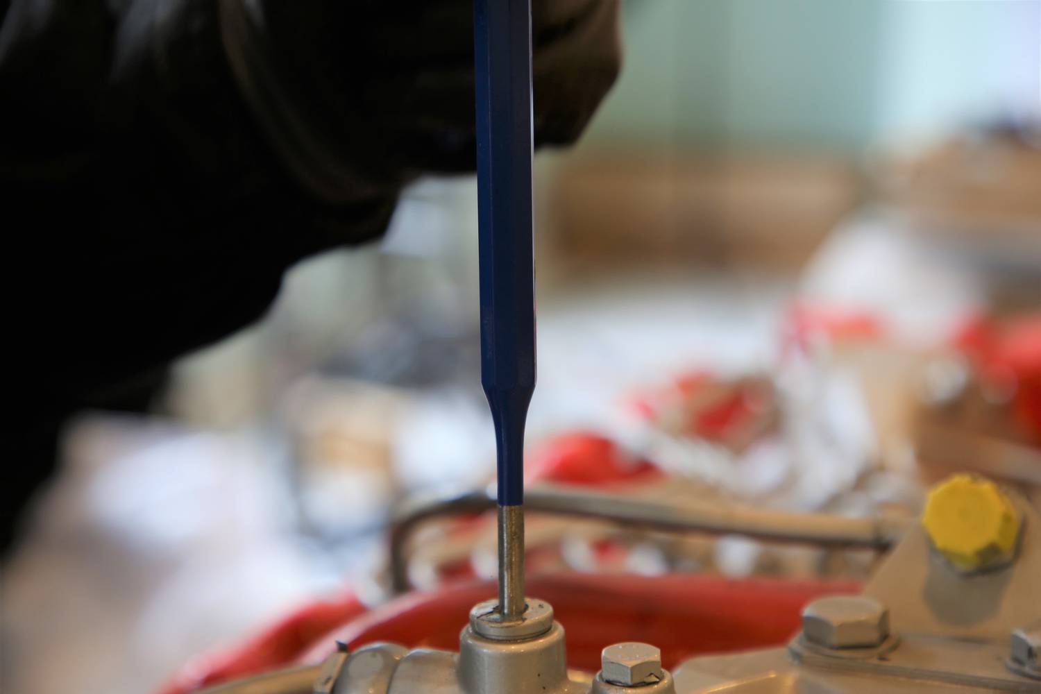 Eine Person repariert oder montiert ein mechanisches Bauteil mit einem blauen Schraubendreher. Im Hintergrund sind Werkstattdetails unscharf.