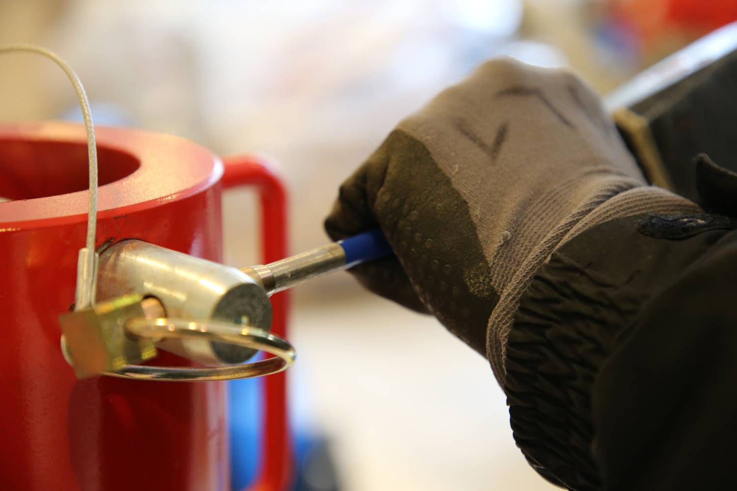Eine behandschuhte Hand bedient ein Werkzeug an einem roten Metallzylinder. Das Werkzeug dient zur Befestigung oder Sicherung.