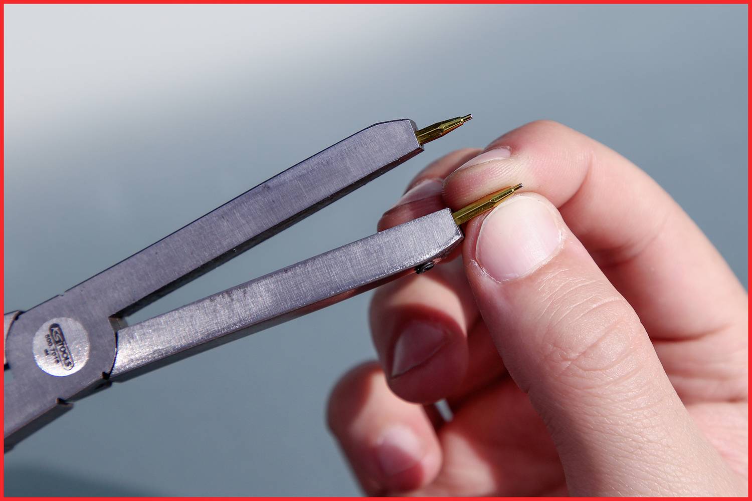 Eine Hand hält eine Pinzette, die einen dünnen Draht greift. Die Pinzette wird von einer zweiten Hand stabilisiert.