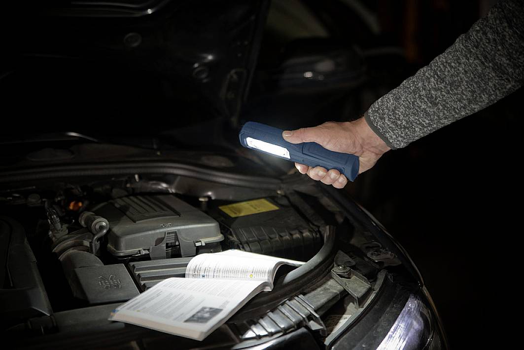 Person hält Taschenlampe über geöffnete Motorhaube eines Autos, beleuchtet Motor und Handbuch.