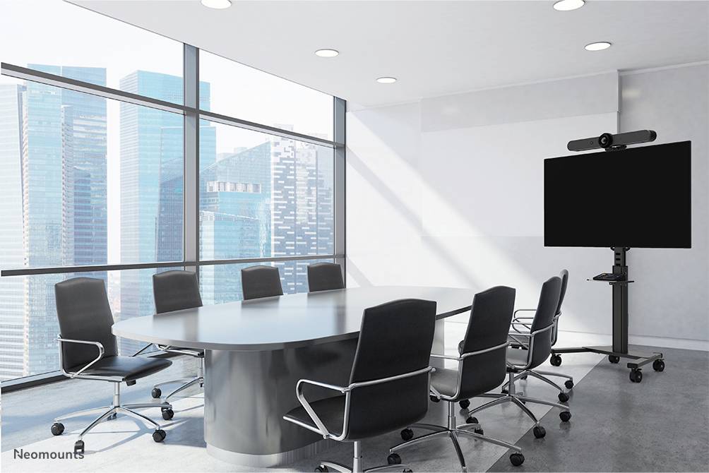 Konferenzraum mit großem ovalen Tisch, acht schwarzen Stühlen und großem Bildschirm auf einem Ständer; Fenster mit Stadtblick im Hintergrund.