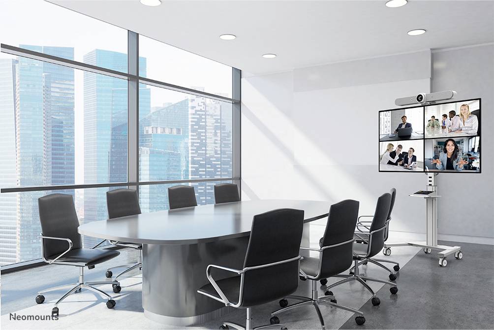 Büro-Konferenzraum mit großem ovalem Tisch und leeren Stühlen. Ein Bildschirm zeigt ein Videomeeting mit mehreren Teilnehmern.