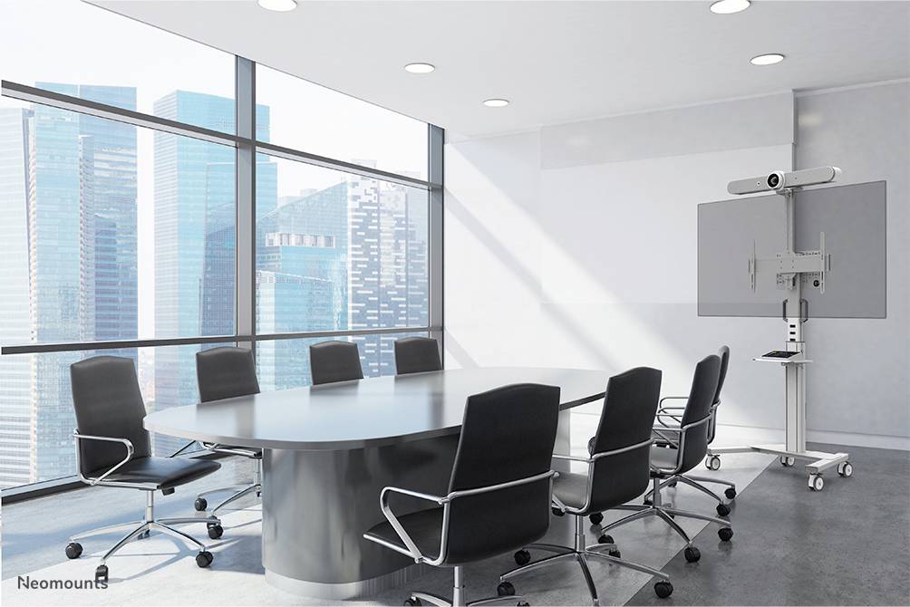 Konferenzraum mit großem ovalen Tisch, umgeben von acht schwarzen Stühlen. Im Hintergrund große Fenster mit Stadtblick, rechts ein Bildschirm.