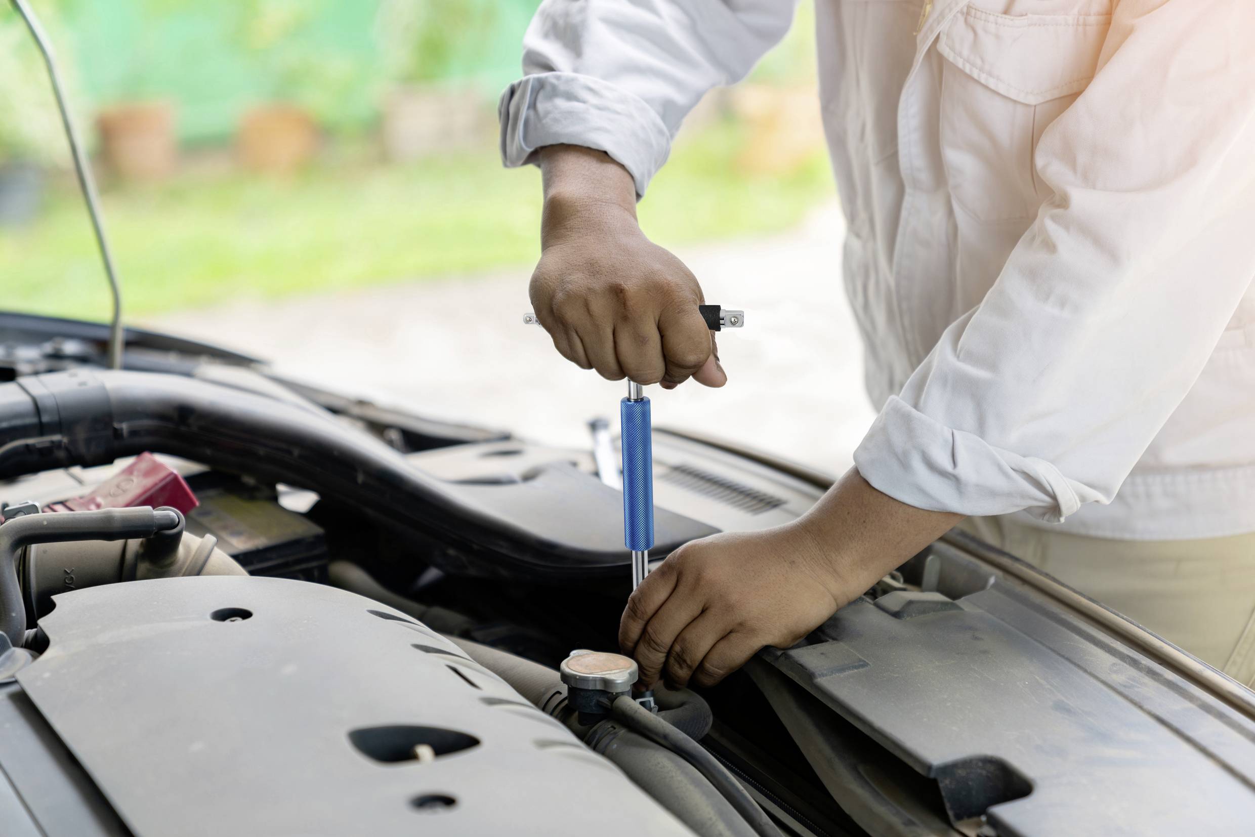 Eine Person in weißem Hemd hält einen Schraubendreher und repariert den Motor eines Autos im Freien.