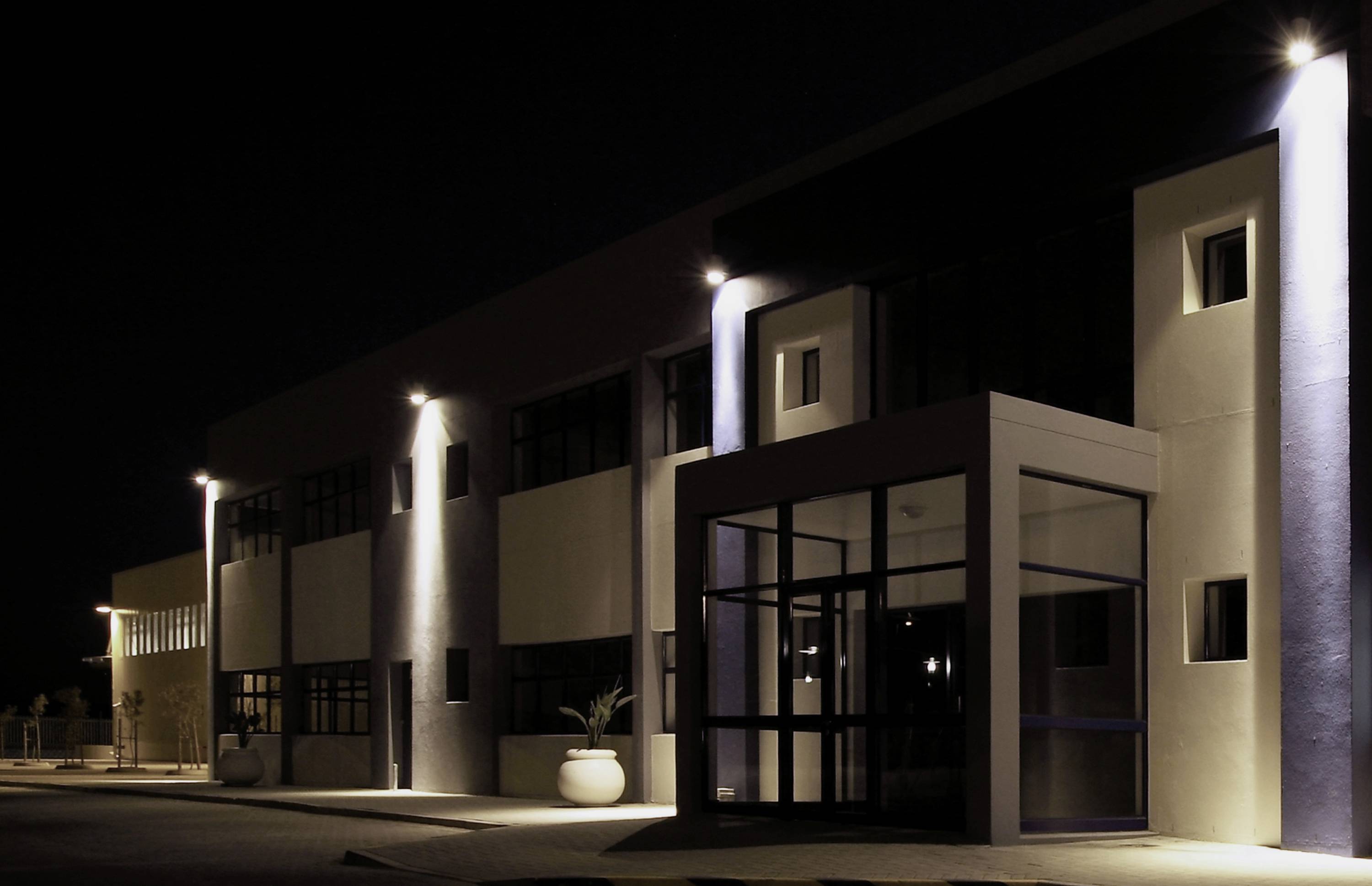 Ein modernes Gebäude bei Nacht, beleuchtet von mehreren Außenleuchten. Die Fassade zeigt große Fenster und einen verglasten Eingangsbereich.