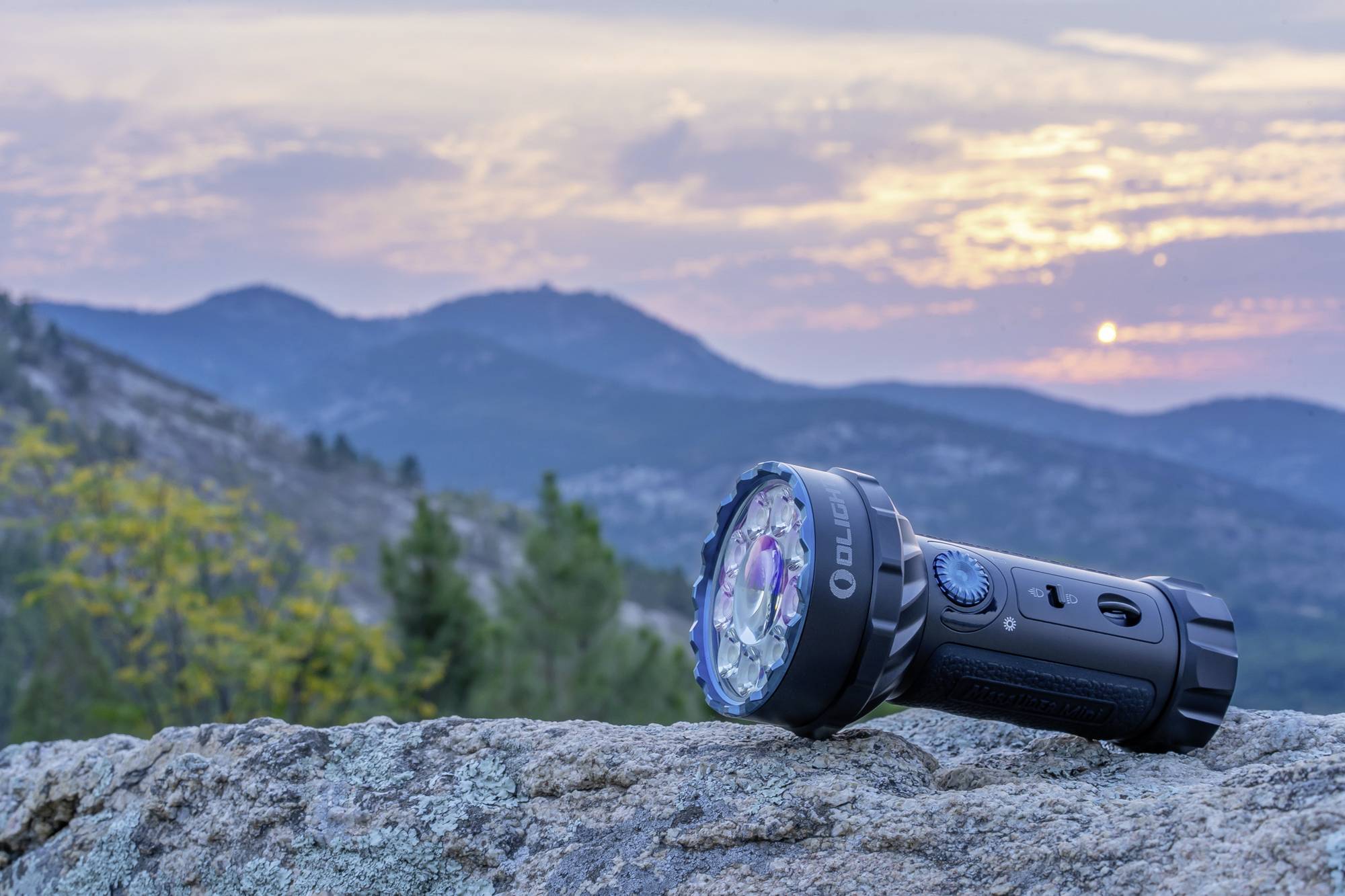 Eine Taschenlampe liegt auf einem Felsen bei Sonnenuntergang in einer Berglandschaft. Der Himmel zeigt Wolken und farbige Lichter.