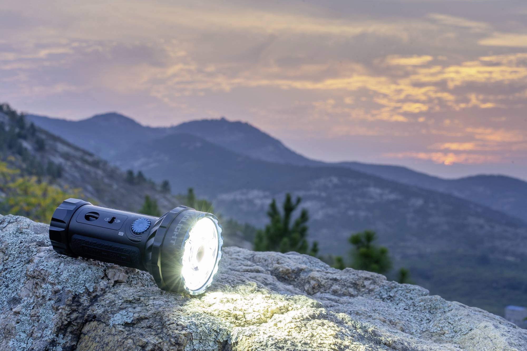 Taschenlampe leuchtet auf einem Stein in der Dämmerung, mit Bergen im Hintergrund. Der Himmel zeigt eine Mischung aus Orange und Lila.