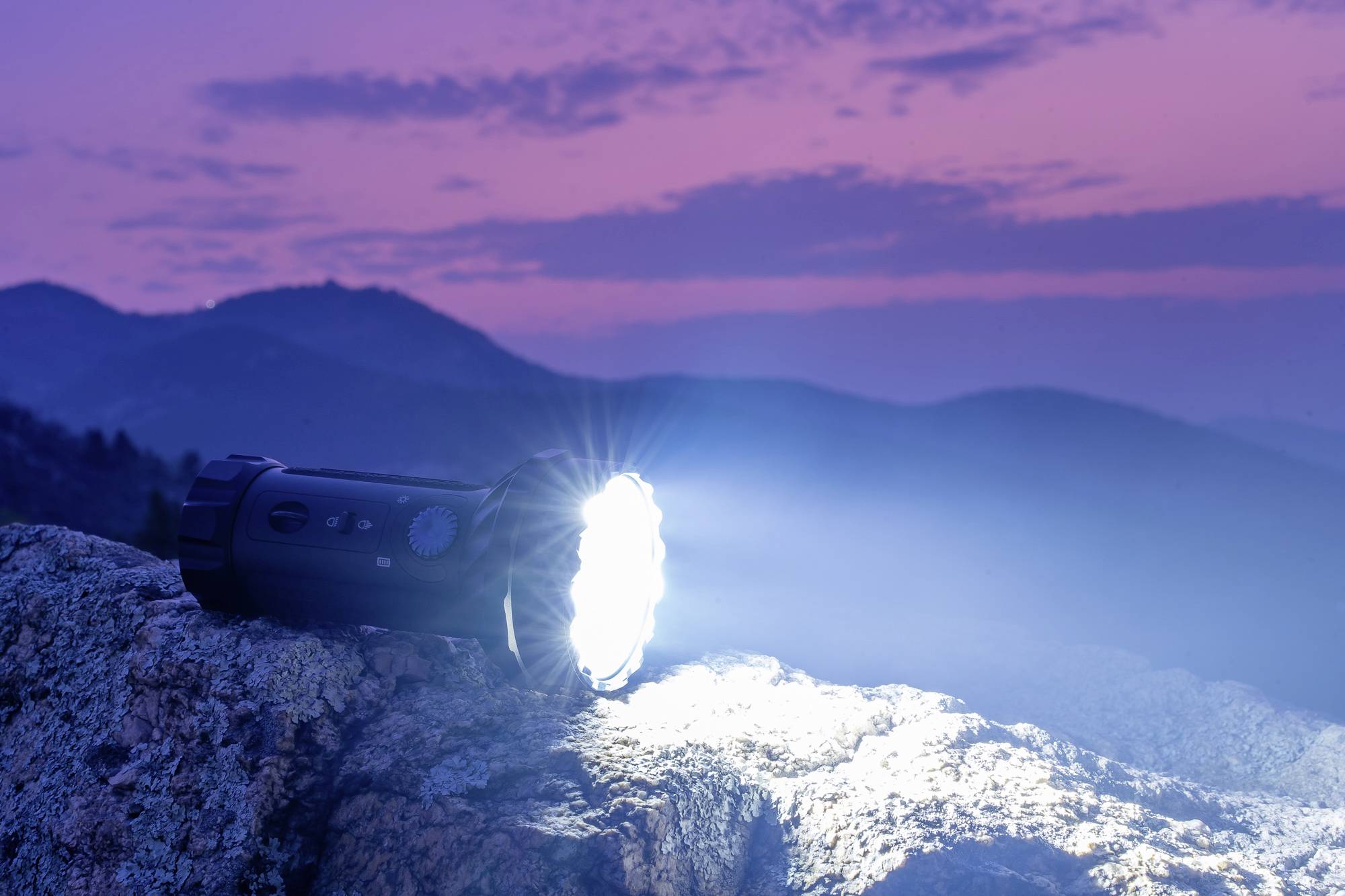 Taschenlampe leuchtet auf einem Felsen in der Dämmerung, umgeben von Hügeln unter einem lila-rosa Himmel.