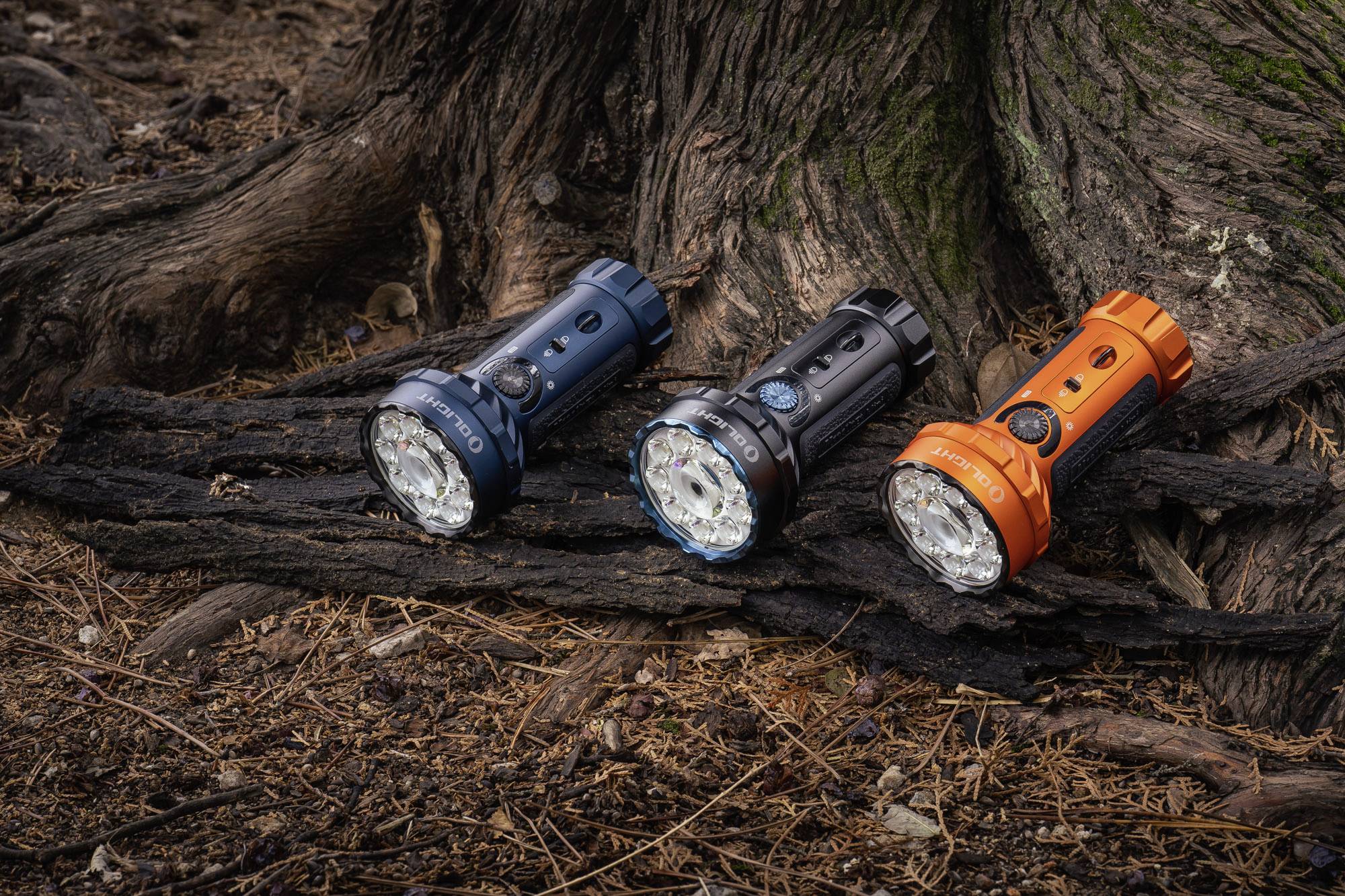 Drei Taschenlampen in Blau, Schwarz und Orange liegen auf einem Baumstamm im Wald, umgeben von Laub und Ästen.