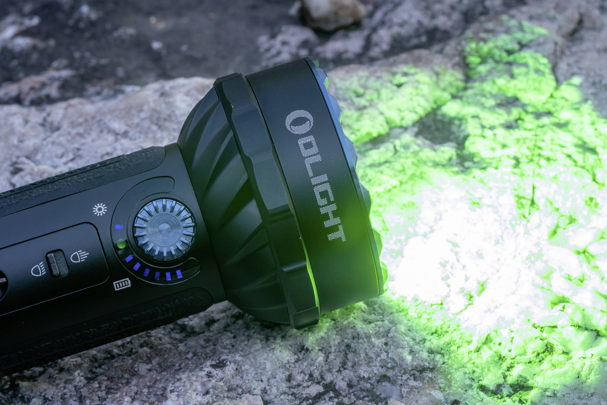 Eine Taschenlampe mit grünem Licht leuchtet auf einen Steinuntergrund. Markenname 'Olight' sichtbar an der Seite der Lampe.