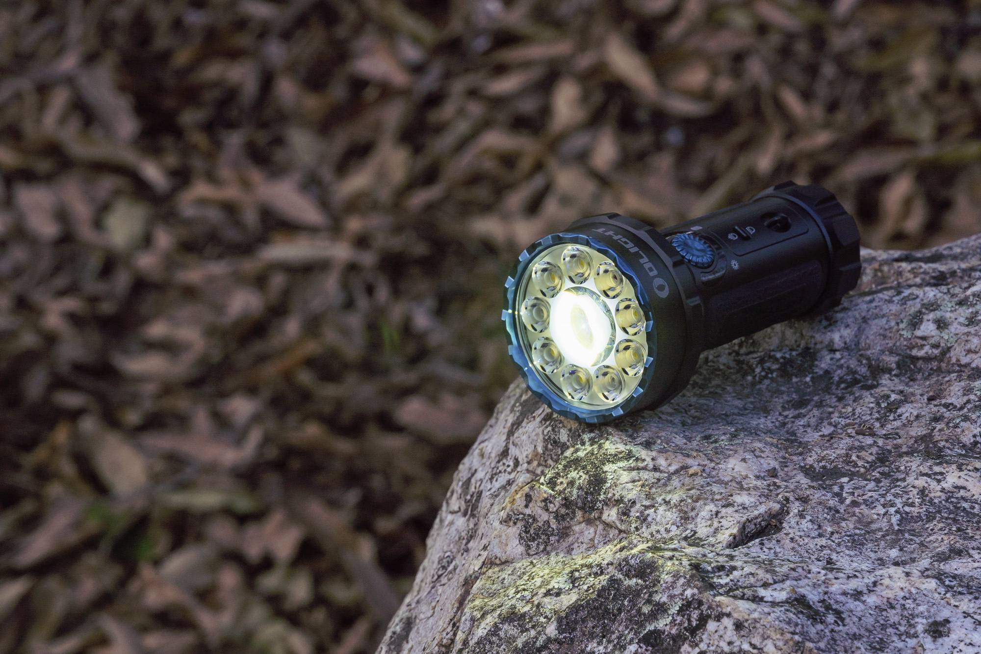 Taschenlampe auf einem Felsen, leuchtend, umgeben von Herbstlaub. Hauptmotiv ist das Licht der Taschenlampe gegen den dunklen Hintergrund.