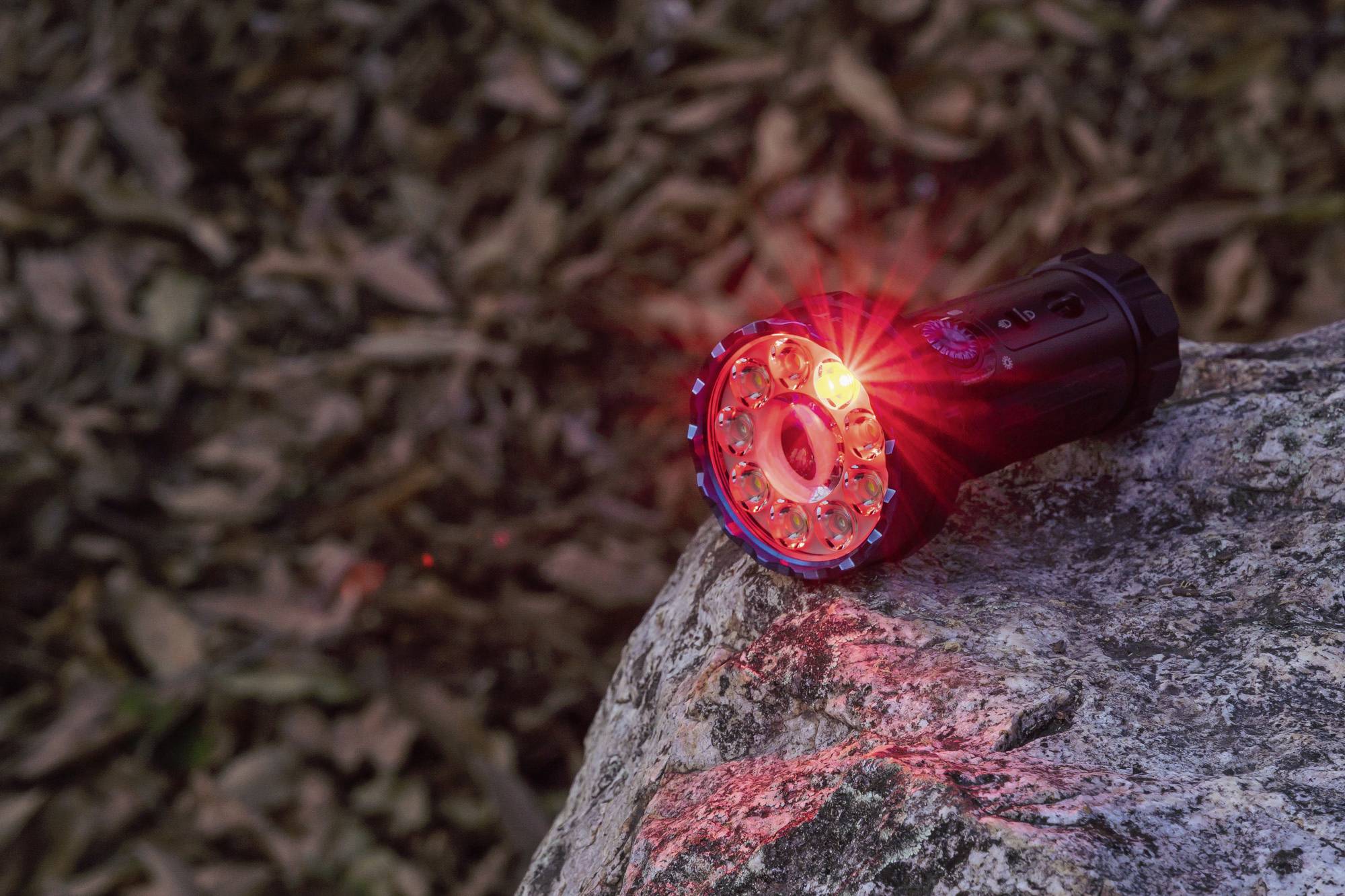 Eine Taschenlampe liegt auf einem Felsen und leuchtet mit rotem Licht. Im Hintergrund sind unscharfe Blätter sichtbar.