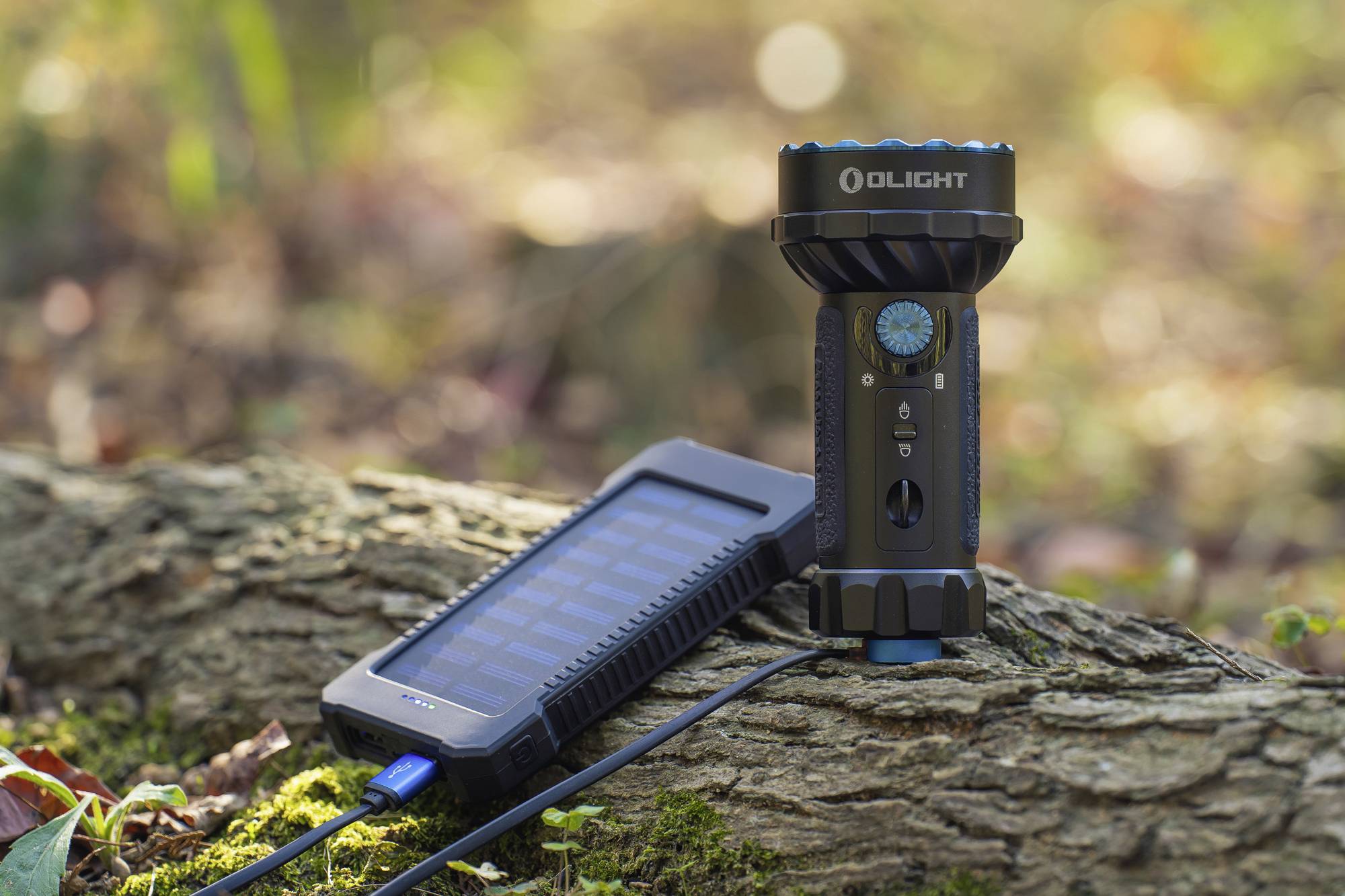 Taschenlampe auf einem Baumstamm im Wald, neben einem Solarladegerät, das mit einem Kabel verbunden ist. Hintergrund aus unscharfem Laub.