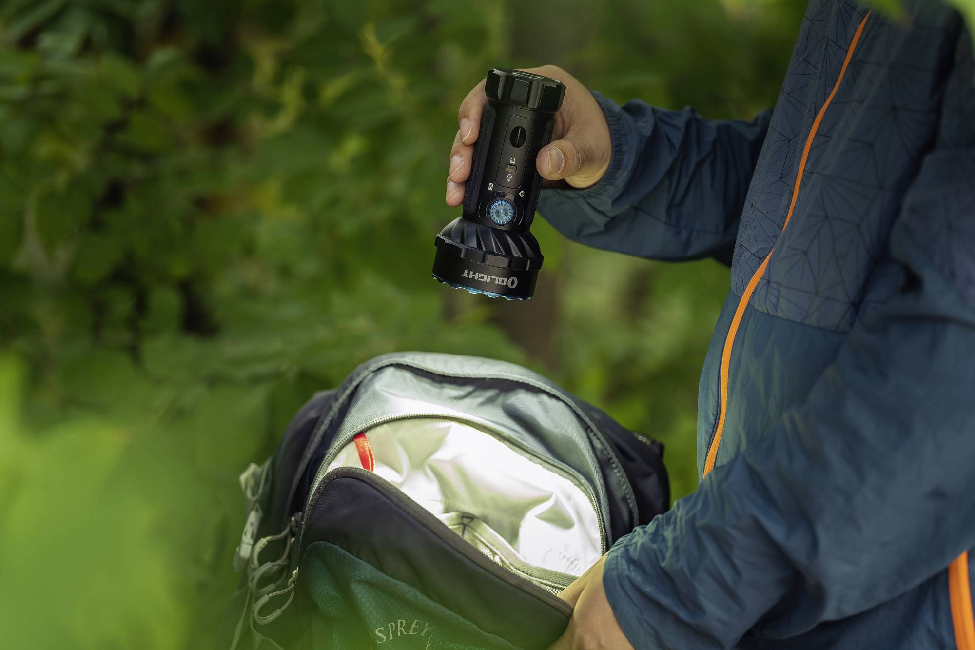 Person hält eine Taschenlampe über einen offenen Rucksack im Wald, bereit für eine Wanderung oder Camping.