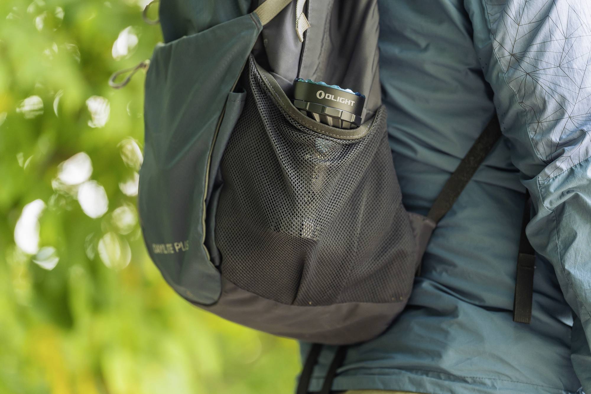 Eine Person trägt einen blauen Rucksack im Freien. In der Seitentasche des Rucksacks steckt eine Taschenlampe.