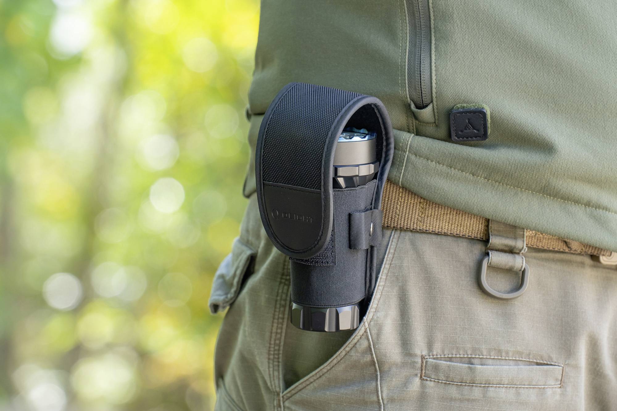 Eine Person trägt ein Fernglas in einer schwarzen Tasche, die am Gürtel befestigt ist. Hintergrund unscharf, zeigt grüne Natur.