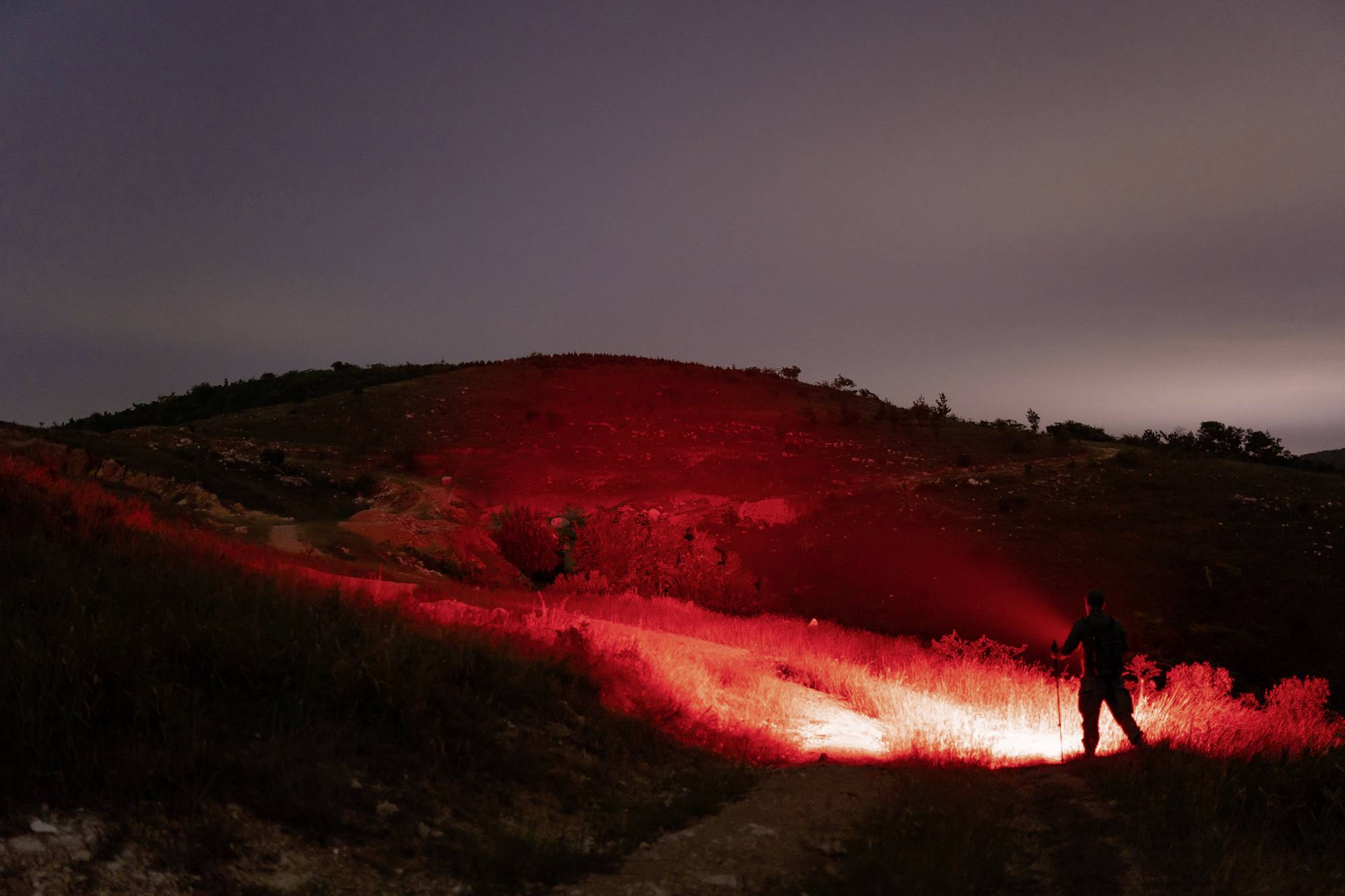 Eine Person steht nachts auf einem Hügel und leuchtet mit einer starken roten Taschenlampe in die Umgebung, die Landschaft ist dunkel.