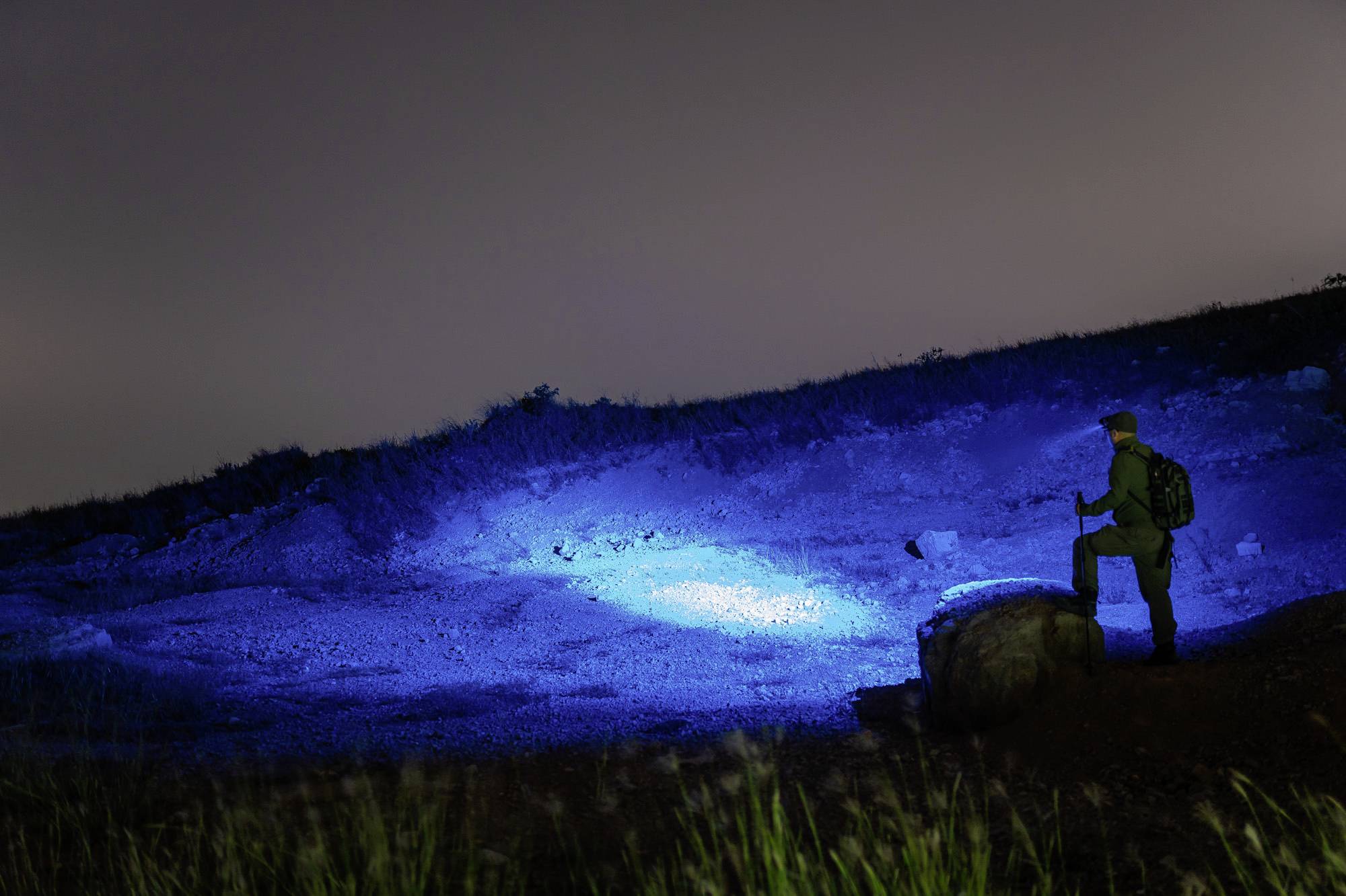 Einsamer Wanderer steht bei Nacht auf einem Hügel, beleuchtet von mysteriösem blauem Licht im Hintergrund.