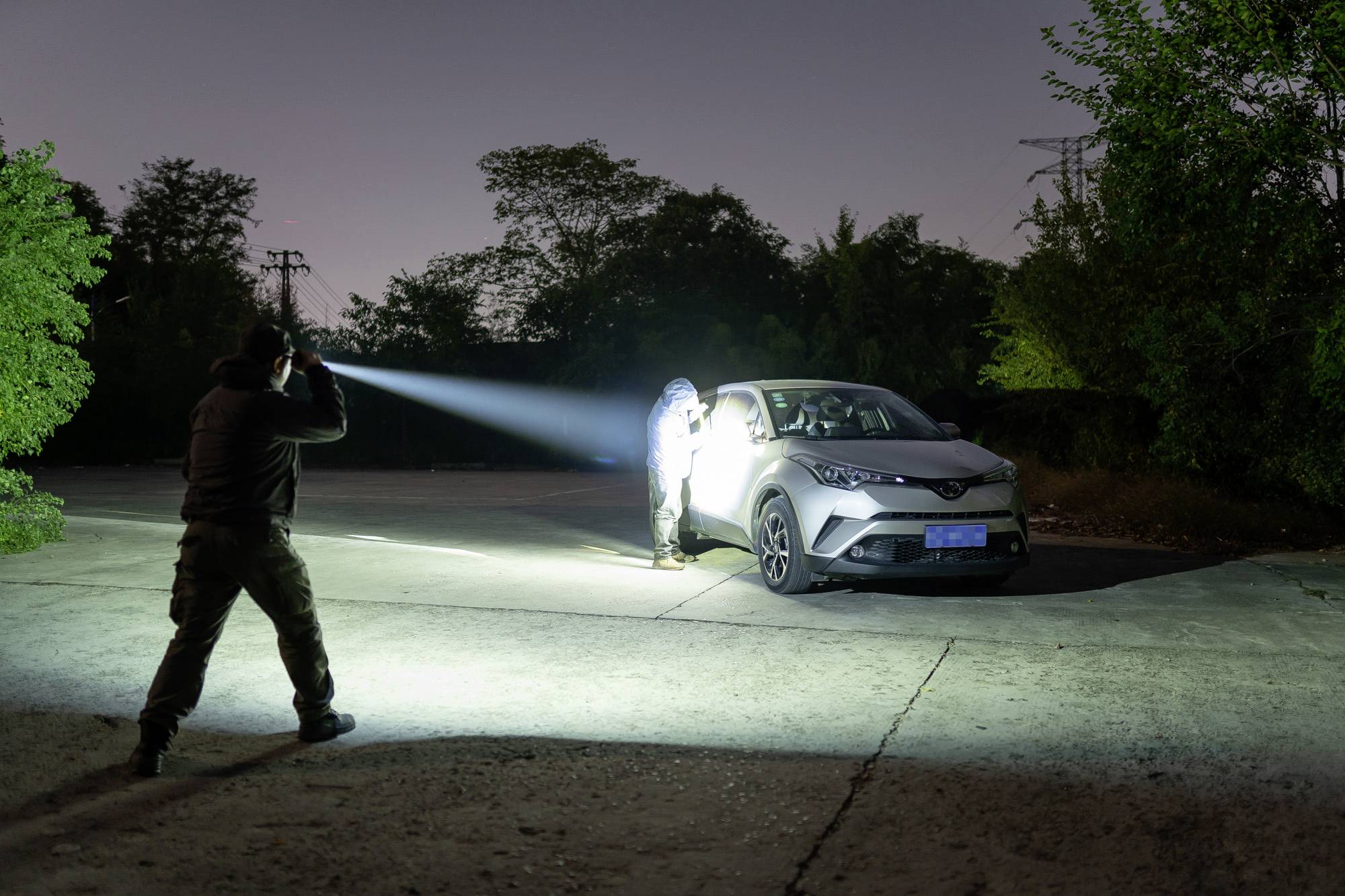Zwei Personen bei Nacht inspizieren ein Auto mit Taschenlampen auf einem leeren Parkplatz, umgeben von Bäumen.