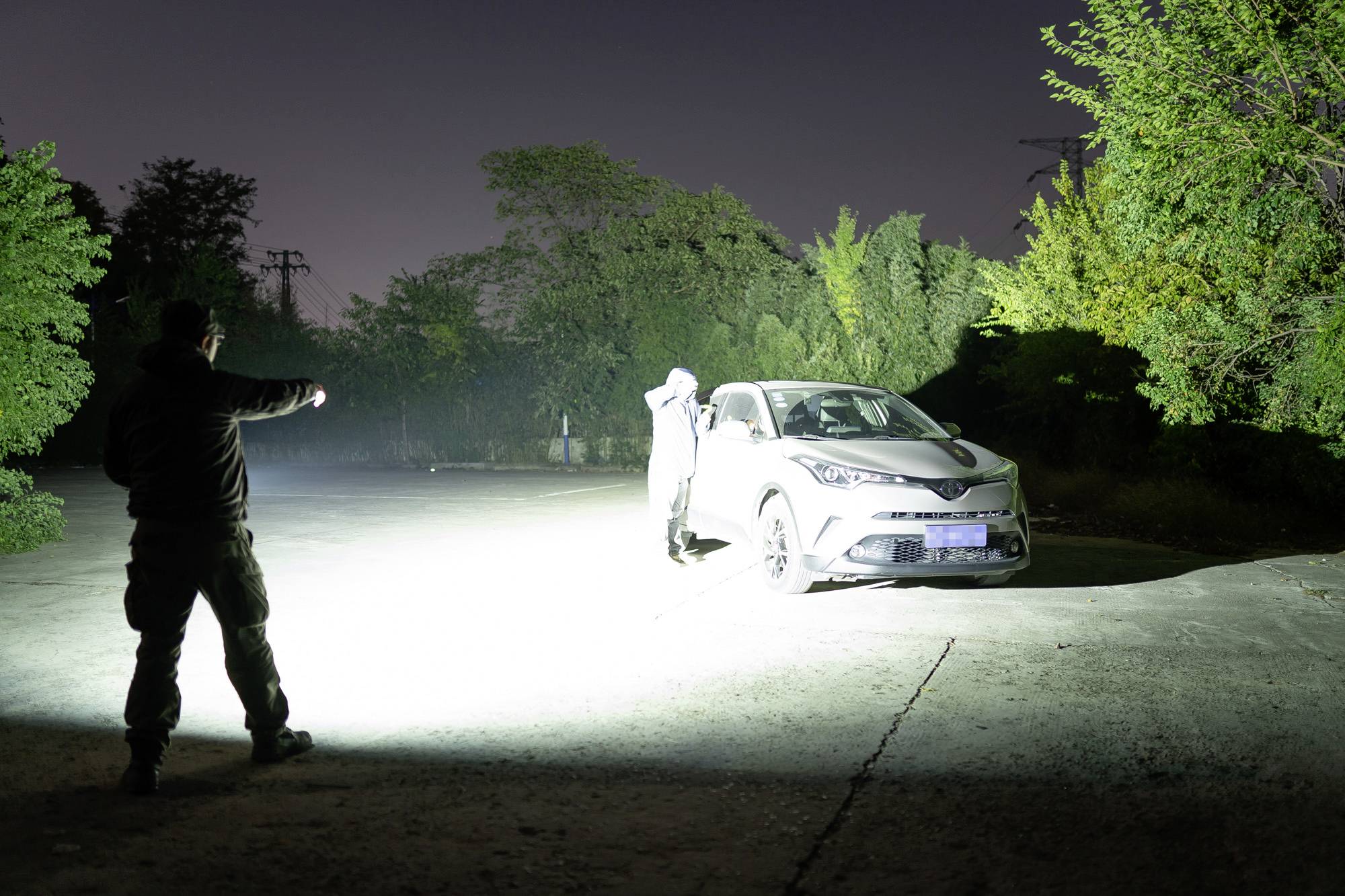 Eine Person leuchtet mit einer starken Taschenlampe auf ein Auto auf einem Parkplatz. Eine andere Person steht am Auto und hebt die Hände.